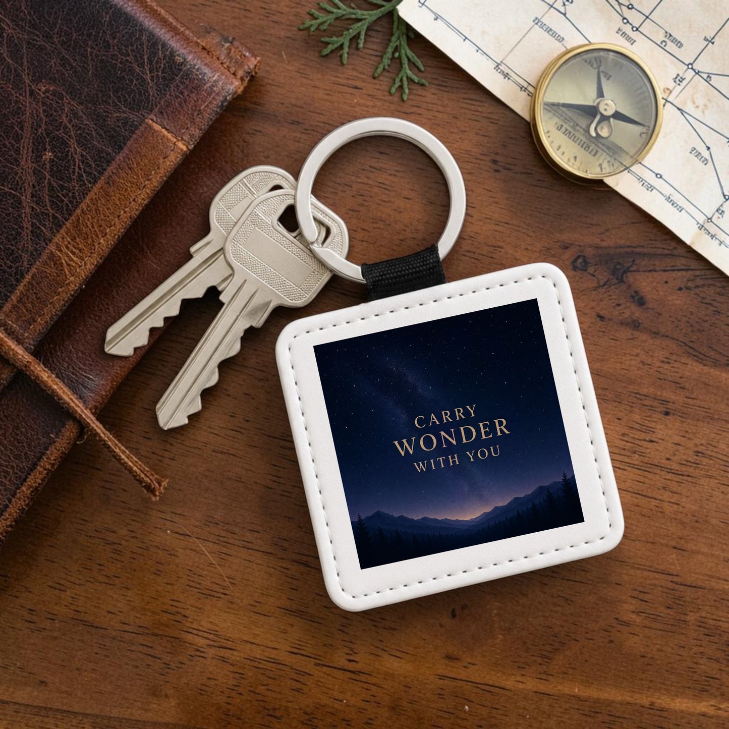 A square Carry Wonder Celestial Keychain from Mirabilia Boutique’s Star & Solstice Collection rests on a wooden table, holding two keys. This decorative keepsake features the phrase “CARRY WONDER WITH YOU” set against a night sky backdrop. Surrounding the keychain are a leather journal, a map, a compass, and a plant.