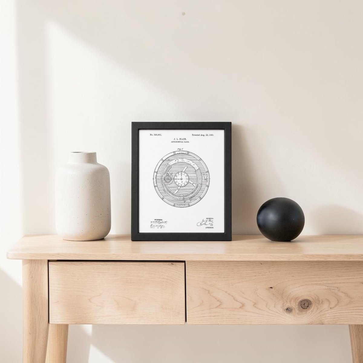 A light wood table displays a white ceramic vase, a black sphere, and the Mirabilia Boutique Astronomical Clock Patent Art Print against an off-white wall. Sunlight creates soft shadows.