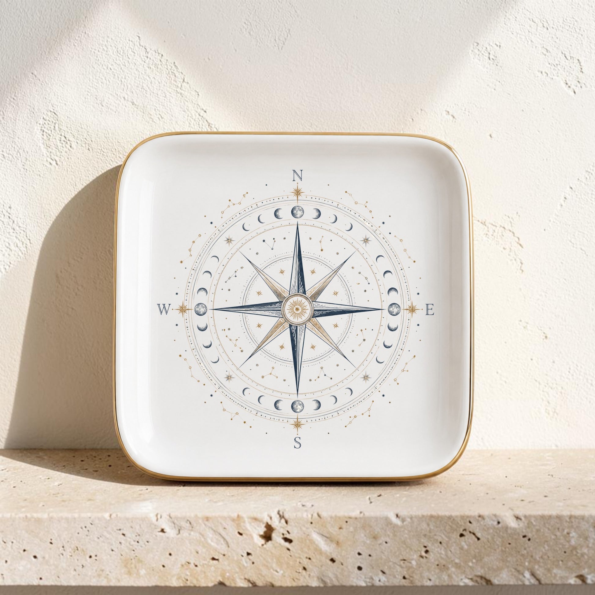 The Celestial Compass Jewelry Tray | Gilded Ceramic by Mirabilia Boutique is a square ceramic plate adorned with a gold rim, showcasing a striking blue compass rose and moon phases design at its center. Resting on a light-colored stone surface against a textured white wall, this constellation-inspired catchall radiates celestial decor charm—making it the perfect stylish accessory for organizing your favorite jewelry pieces.