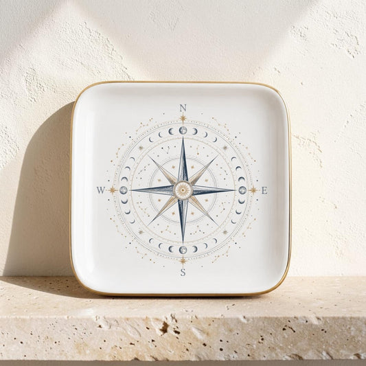 The Celestial Compass Jewelry Tray | Gilded Ceramic by Mirabilia Boutique is a square ceramic plate adorned with a gold rim, showcasing a striking blue compass rose and moon phases design at its center. Resting on a light-colored stone surface against a textured white wall, this constellation-inspired catchall radiates celestial decor charm—making it the perfect stylish accessory for organizing your favorite jewelry pieces.
