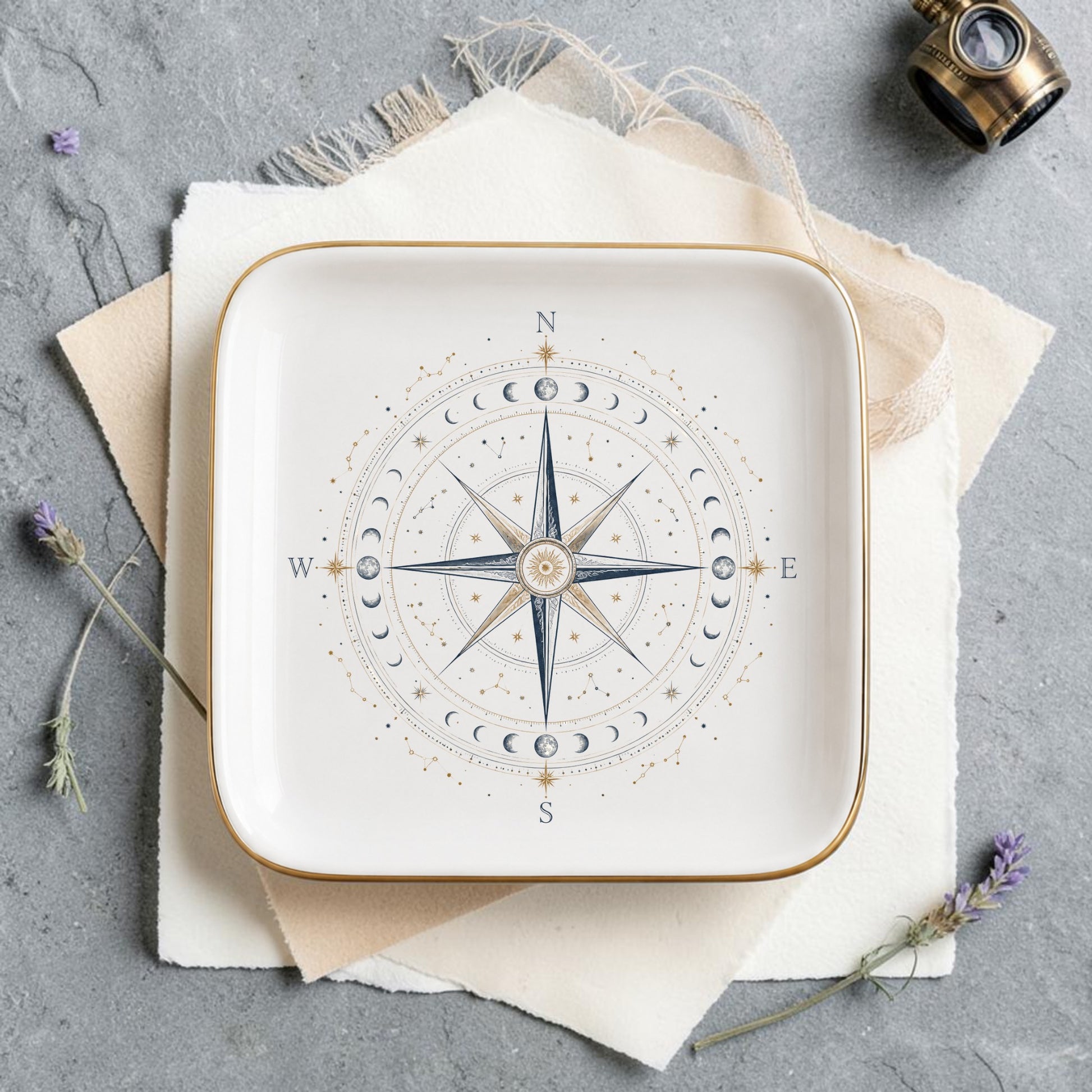 The Celestial Compass Jewelry Tray | Gilded Ceramic by Mirabilia Boutique features a square plate adorned with a compass rose and moon phases, elegantly displayed on layered paper atop a gray surface, accented by lavender sprigs and a vintage brass compass—an enchanting celestial decor piece or constellation catchall for your space.