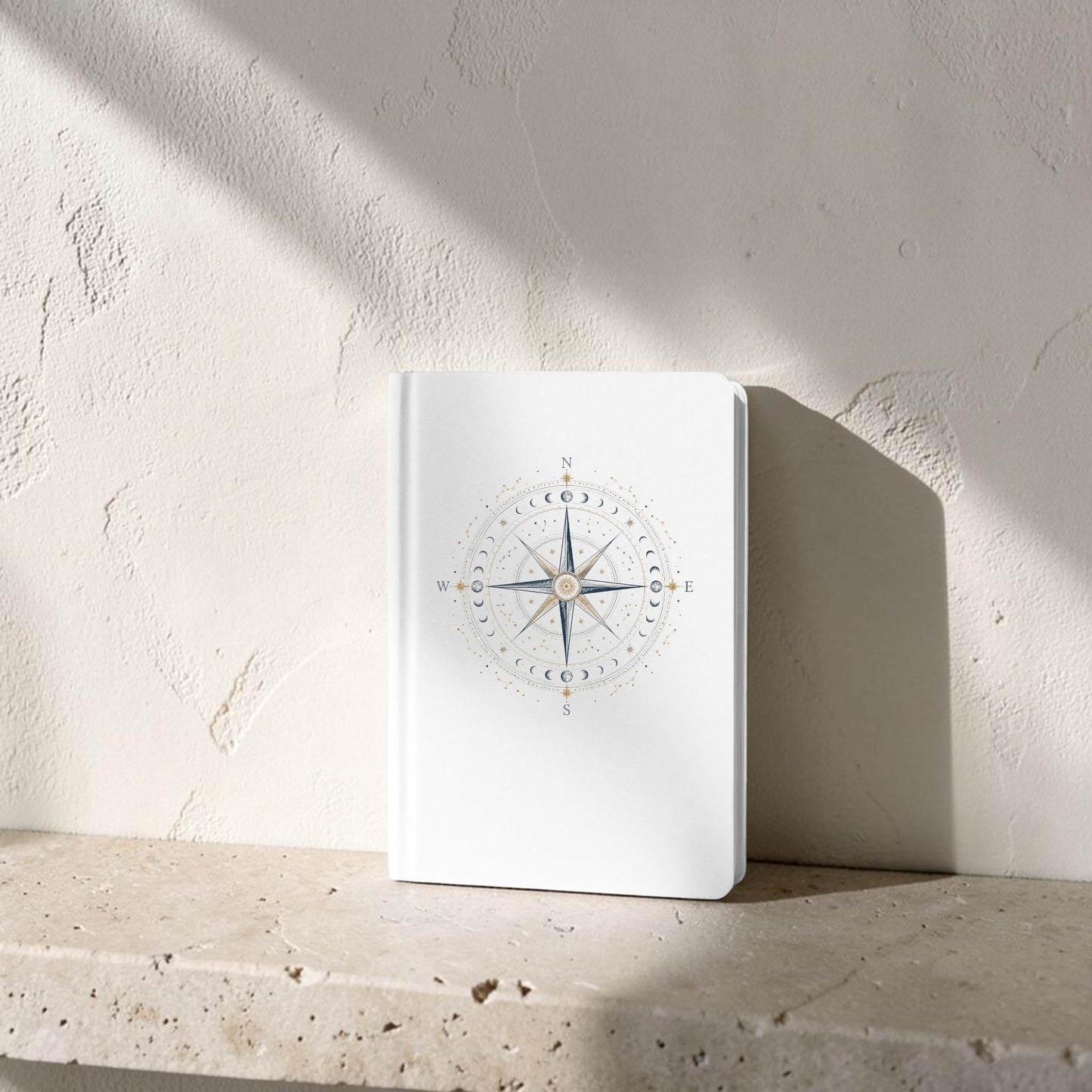 A Celestial Compass Journal | Moon Phase Notebook by Mirabilia Boutique, featuring a compass rose design on its white cover, stands upright on a stone ledge against a textured wall bathed in sunlight.