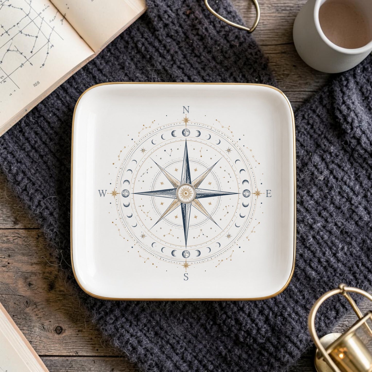Add a touch of cosmic elegance to your space with the Celestial Compass Jewelry Tray | Gilded Ceramic by Mirabilia Boutique—a square plate adorned with an intricate compass rose and moon phases design. Resting on a knitted gray cloth and surrounded by a coffee cup, a book of star charts, and a brass accent atop a wooden surface, this tray is the perfect celestial décor accent or constellation catchall for your home.