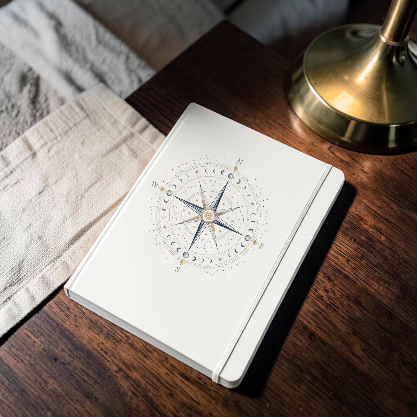 The Celestial Compass Journal | Moon Phase Notebook by Mirabilia Boutique, featuring an intricate compass rose and moon phase design on a white cover, rests on a wooden nightstand beside a gold lamp and a folded beige cloth.