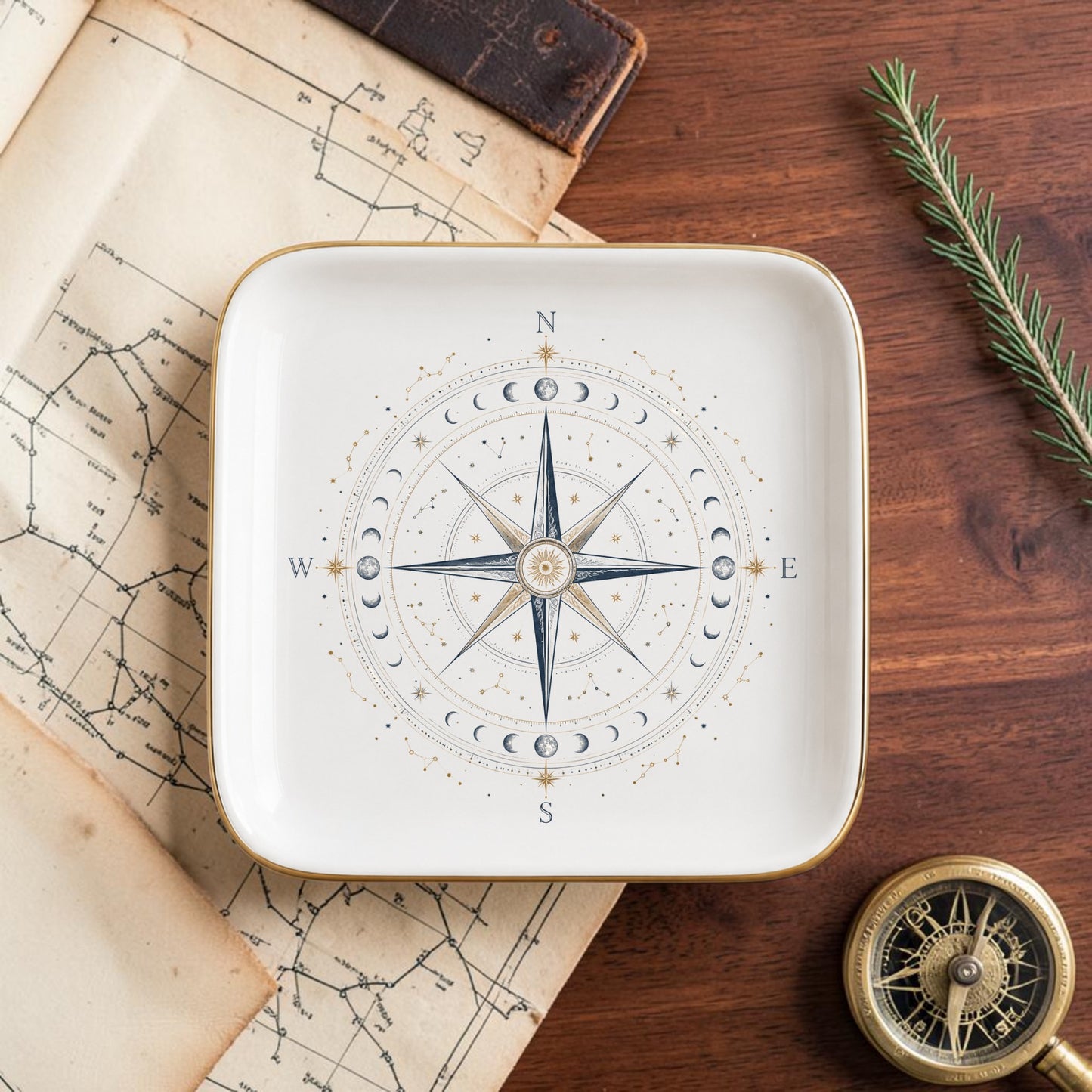 The Celestial Compass Jewelry Tray | Gilded Ceramic by Mirabilia Boutique, adorned with a compass rose and moon phases design, rests on a wooden table alongside a map, compass, leather-bound book, and a sprig of greenery—an elegant way to infuse your space with celestial decor.