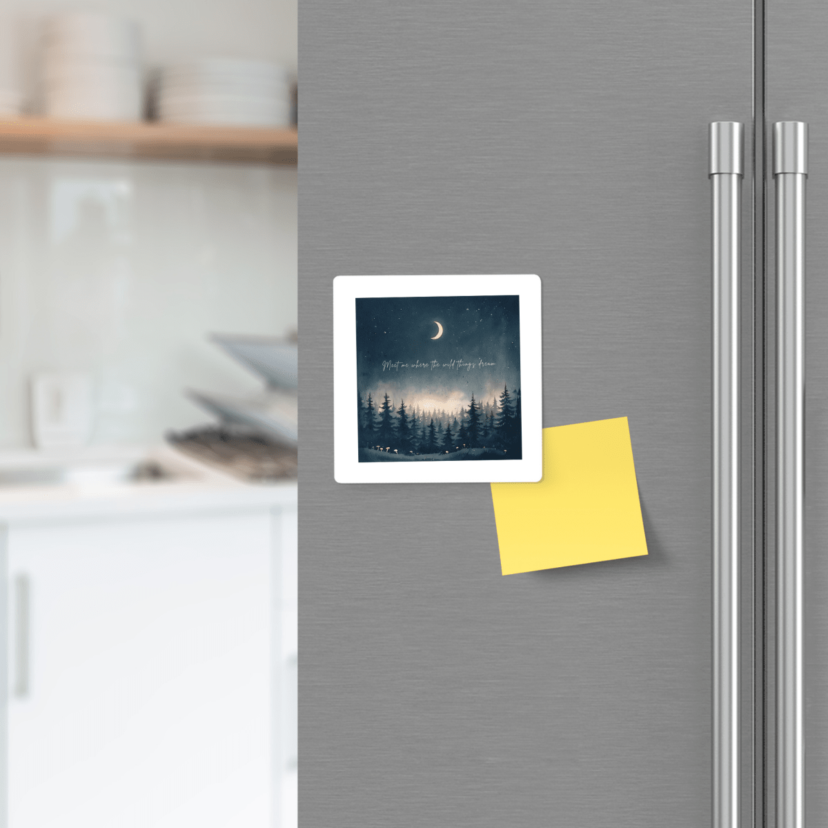 A square photo of a moonlit forest and a yellow sticky note are held to a stainless steel refrigerator in a modern kitchen by the Meet Me Where the Wild Things Dream Magnet from Mirabilia Boutique, adding a touch of celestial decor with its nature-inspired woodland design.