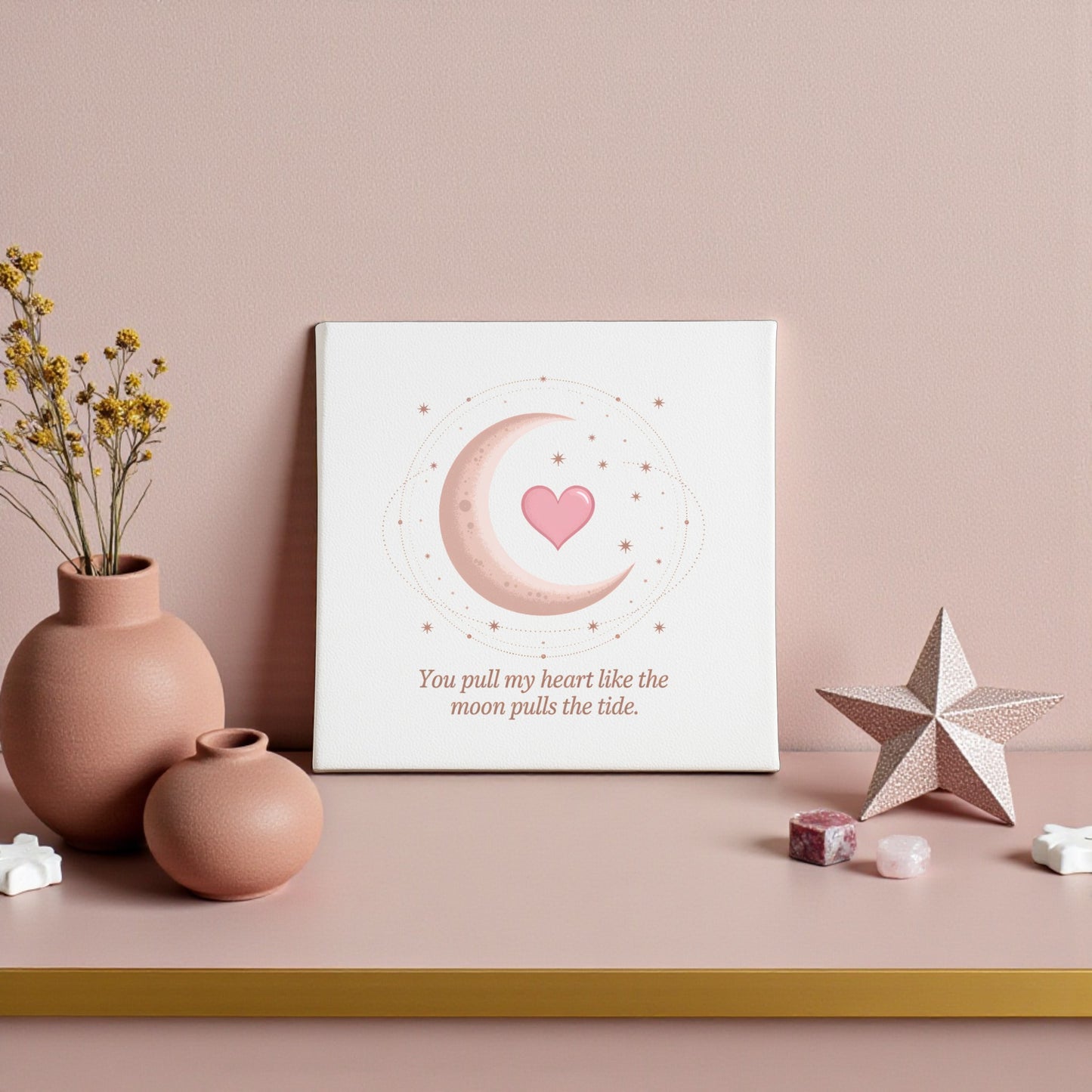 The Moon Pulls the Tide Love Quote Canvas by Mirabilia Boutique features a crescent moon, stars, and pink heart, displayed on a pink shelf with ceramics and crystals. The art reads, “You pull my heart like the moon pulls the tide.”.