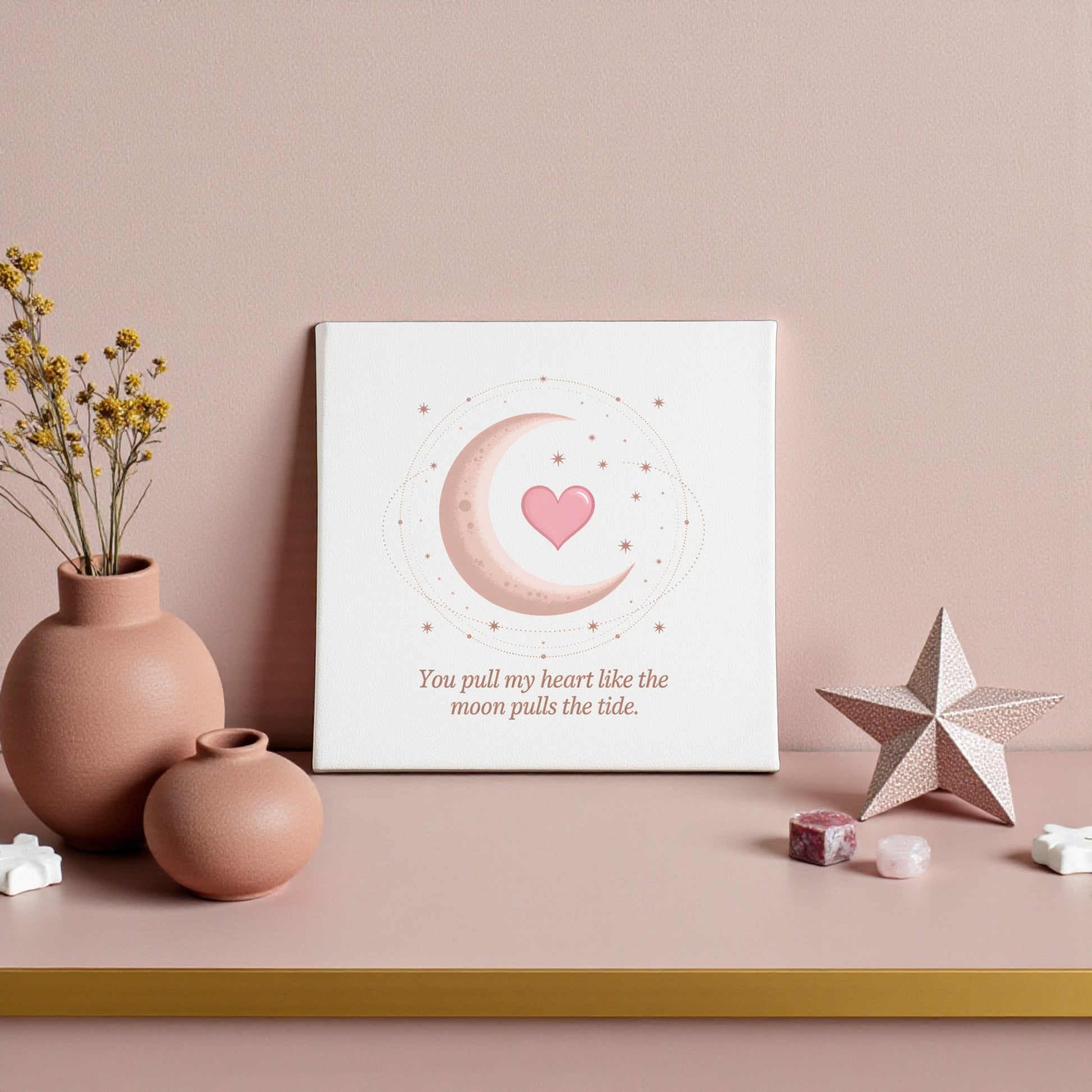 The Moon Pulls the Tide Love Quote Canvas by Mirabilia Boutique features a crescent moon, stars, and pink heart, displayed on a pink shelf with ceramics and crystals. The art reads, “You pull my heart like the moon pulls the tide.”.