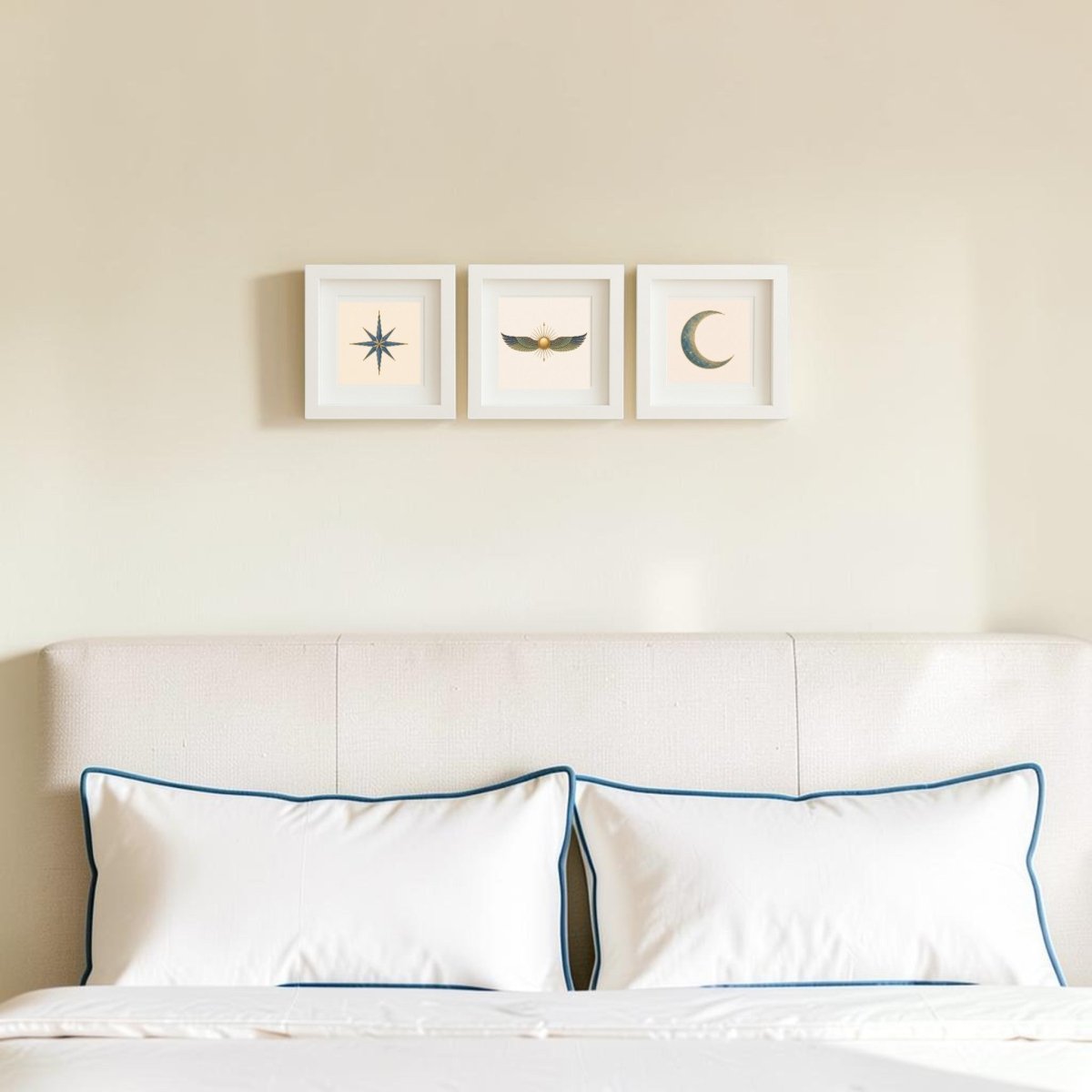 A neatly made bed with two white pillows sits beneath three pieces from the “Celestial Relics of the Nile” collection by Mirabilia Boutique, each displayed in a shadowbox frame and featuring a star, wings, and a crescent moon on a light beige wall.
