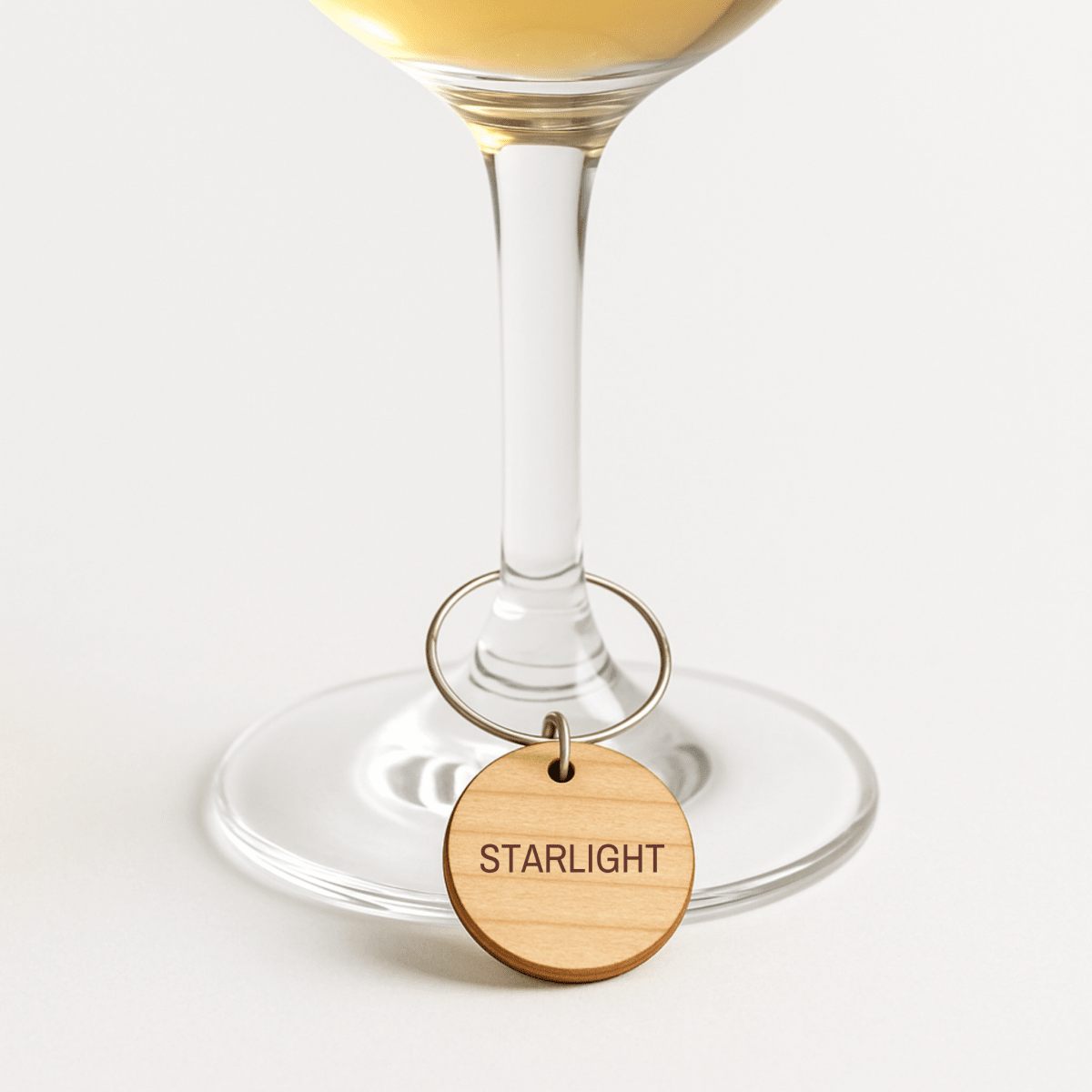 A wine glass adorned with a celestial-themed, laser-engraved wooden charm from the Mirabilia Boutique Star & Solstice Engraved Wood Wine Charms Set of 8, shown hanging from the stem and labeled “STARLIGHT,” photographed on a white background.