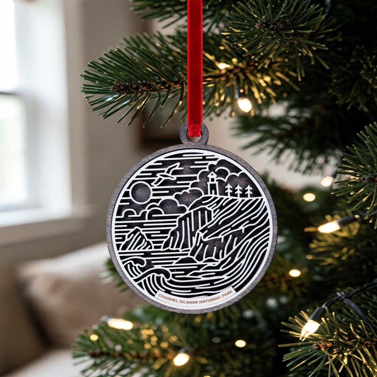 The Channel Islands National Park Christmas Ornament by Mirabilia Boutique—a round black and white wooden ornament showcasing a line art landscape of the national park—hangs from a red ribbon on a Christmas tree adorned with white lights. In the softly lit background, a window and couch can be seen.