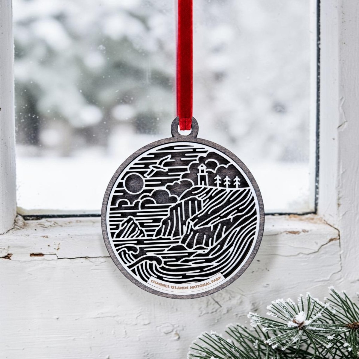 The Channel Islands National Park Christmas Ornament by Mirabilia Boutique features a round metal design with an etched landscape scene, hanging from a red ribbon in front of a snowy window. Snow-dusted pine needles appear in the bottom right corner.
