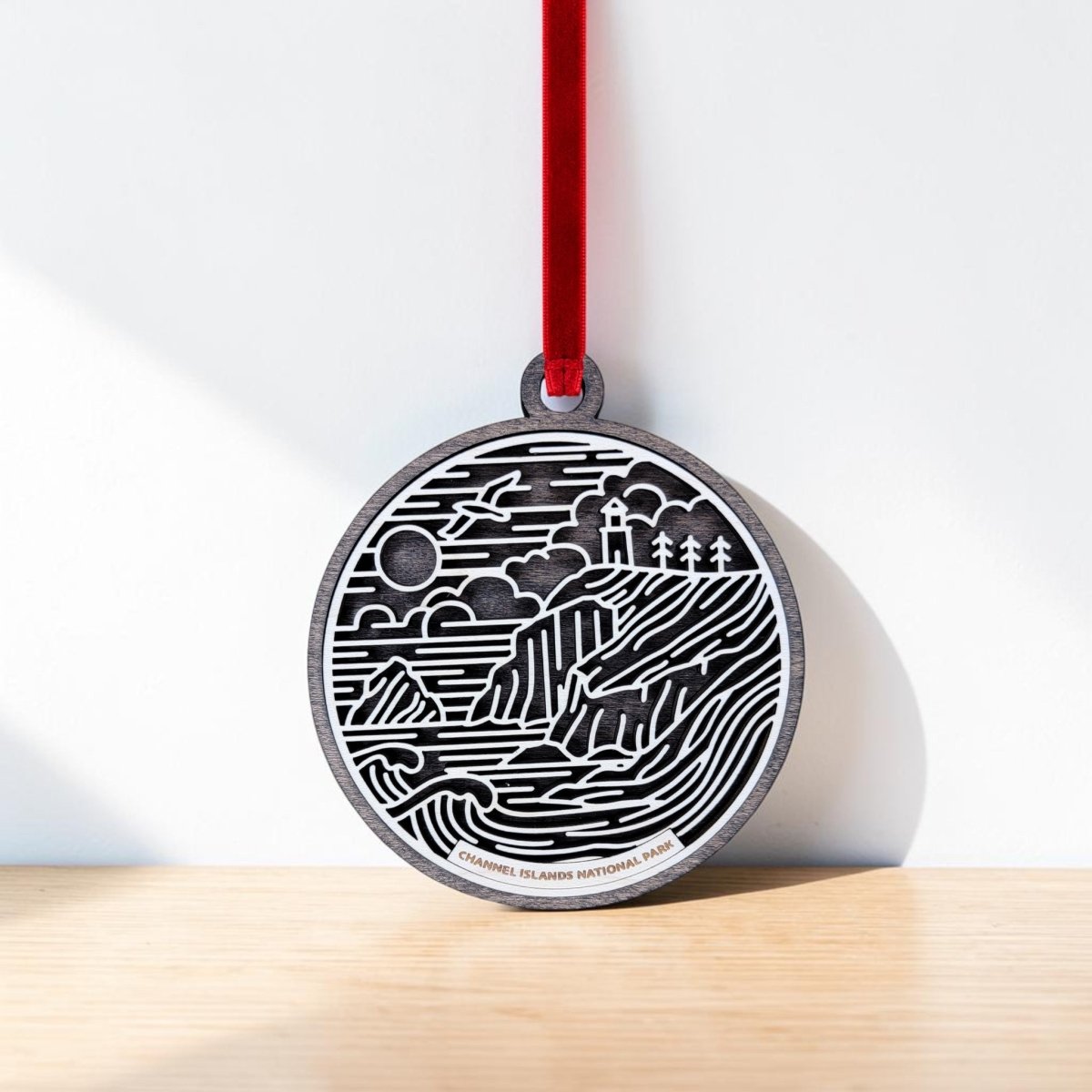 The Channel Islands National Park Christmas Ornament by Mirabilia Boutique is a round wooden decoration featuring a black and white line art design of mountains, ocean waves, birds, and trees. It hangs from a red ribbon against a white wall and wooden surface.