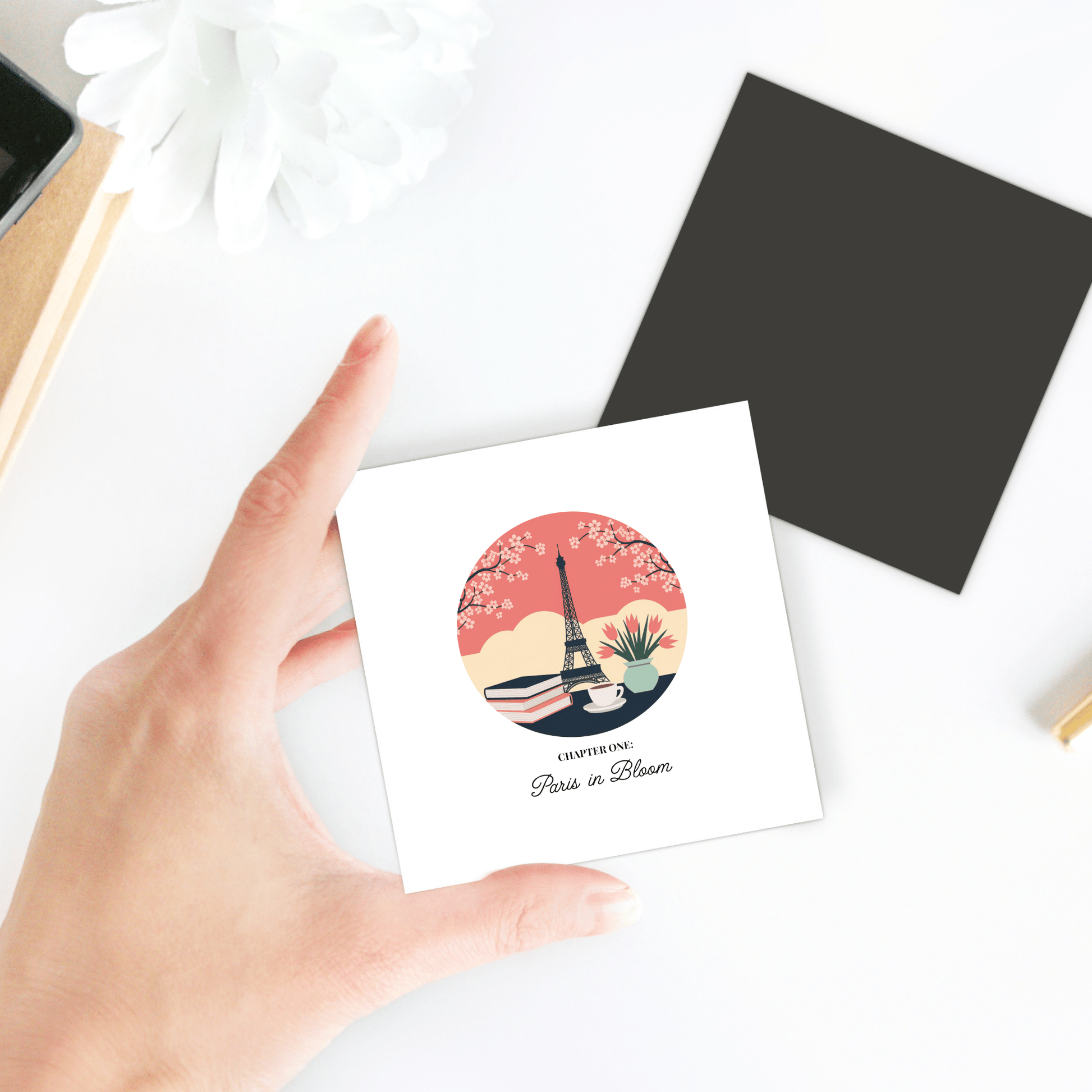 A hand holds the Paris in Bloom Magnet by Mirabilia Boutique, featuring a floral Eiffel Tower illustration and “CAPTURED: Paris in Bloom”—a perfect literary Paris gift for book lovers. A black envelope and white flower sit nearby.