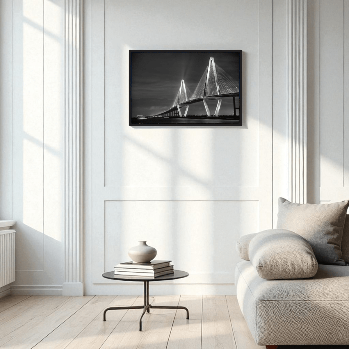 A modern, minimalist living room with white walls, a light sofa, a round black coffee table with a vase, and the Mirabilia Boutique Ravenel Bridge Black & White Fine Art Print | Framed Art Print displayed on the wall.