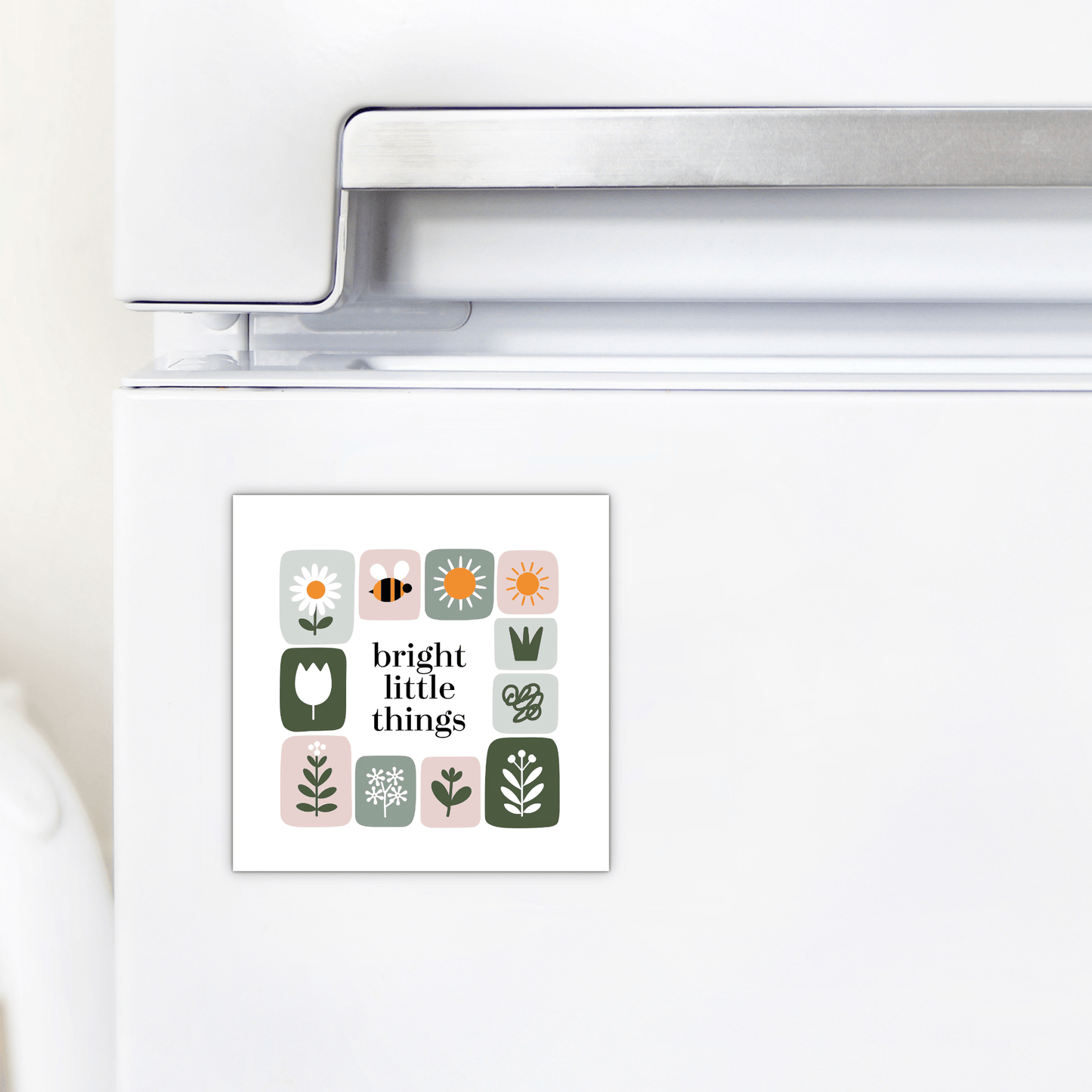 A white refrigerator is decorated with the Mirabilia Boutique Bright Little Things Floral Grid Magnet, featuring simple floral and nature illustrations, a bee, and the words "bright little things" in the center.