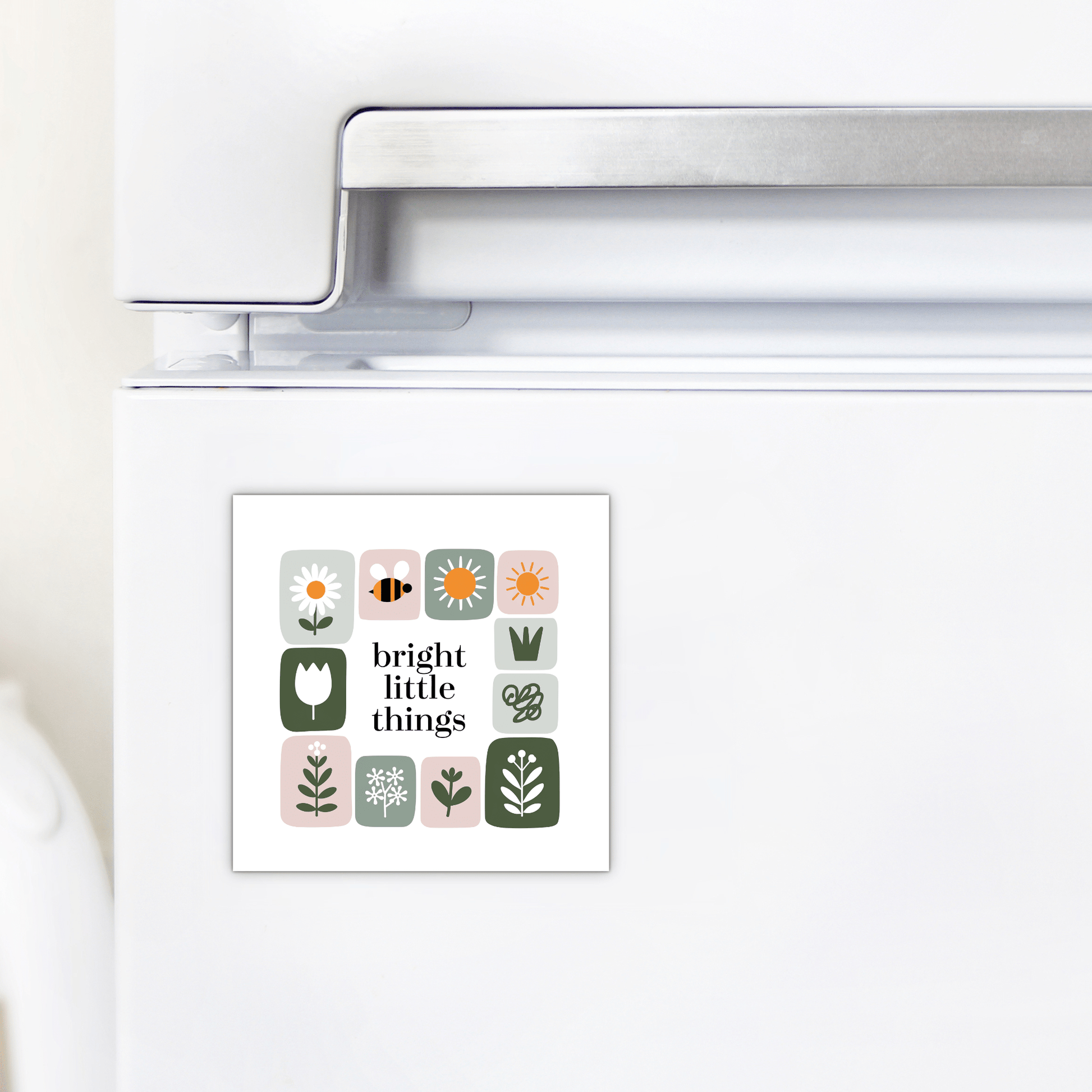 A white refrigerator is decorated with the Mirabilia Boutique Bright Little Things Floral Grid Magnet, featuring simple floral and nature illustrations, a bee, and the words "bright little things" in the center.