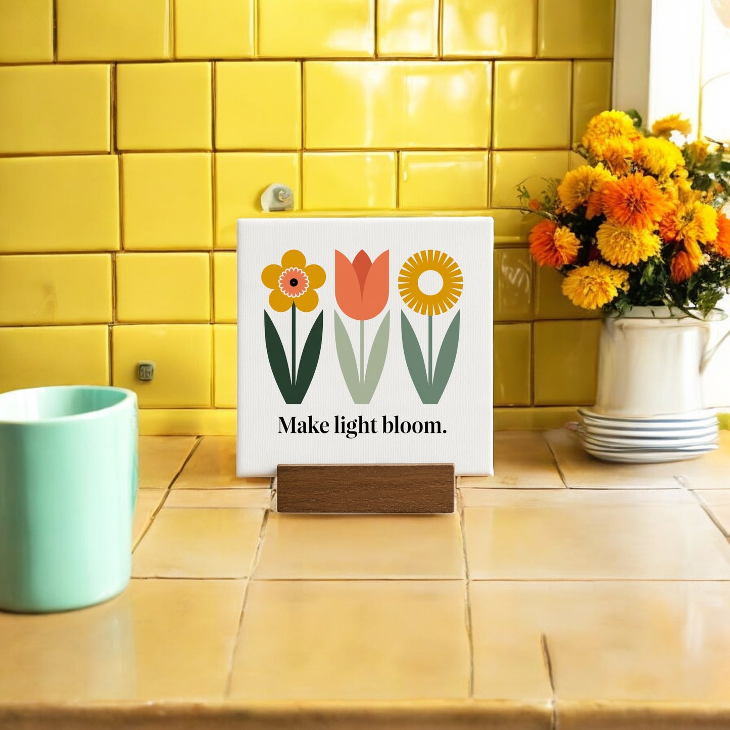 The Make Light Bloom Mini Canvas Sign with Wood Base by Mirabilia Boutique features colorful flower art and an inspiring message, displayed on a kitchen counter with yellow tiles, orange flowers, and a green mug—perfect daily affirmation decor.