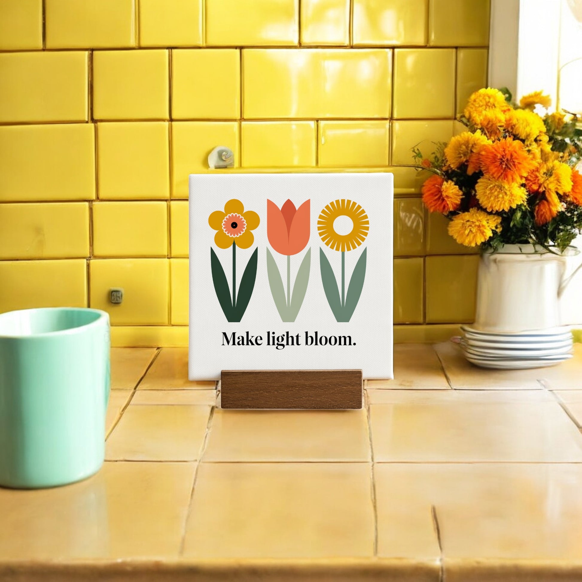 The Make Light Bloom Mini Canvas Sign with Wood Base by Mirabilia Boutique features colorful flower art and an inspiring message, displayed on a kitchen counter with yellow tiles, orange flowers, and a green mug—perfect daily affirmation decor.