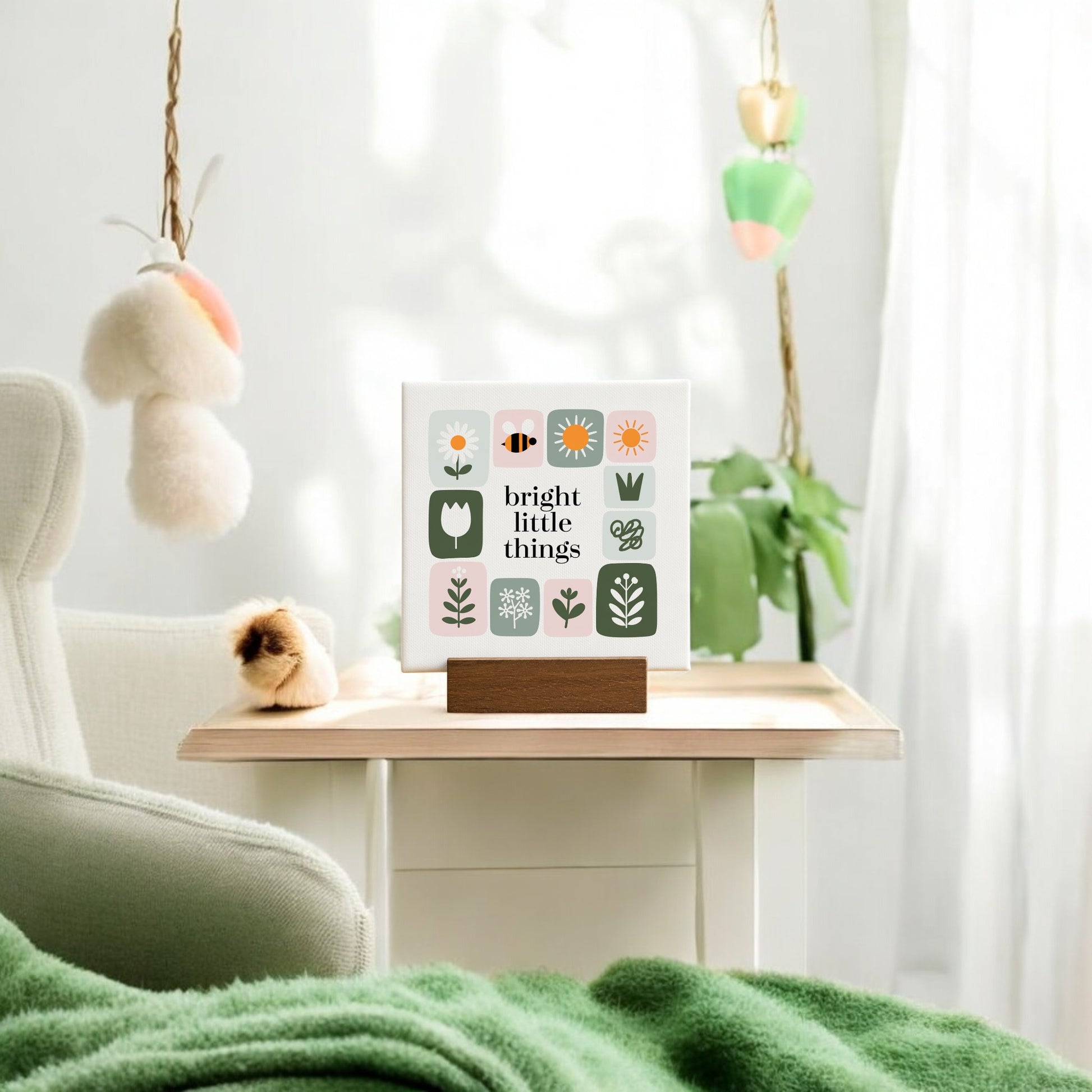 A cozy room features a green blanket, white chair, and small wooden table displaying the Mirabilia Boutique Bright Little Things Mini Canvas Sign with floral art. Handcrafted plush decor hangs nearby as sunlight filters through sheer curtains.
