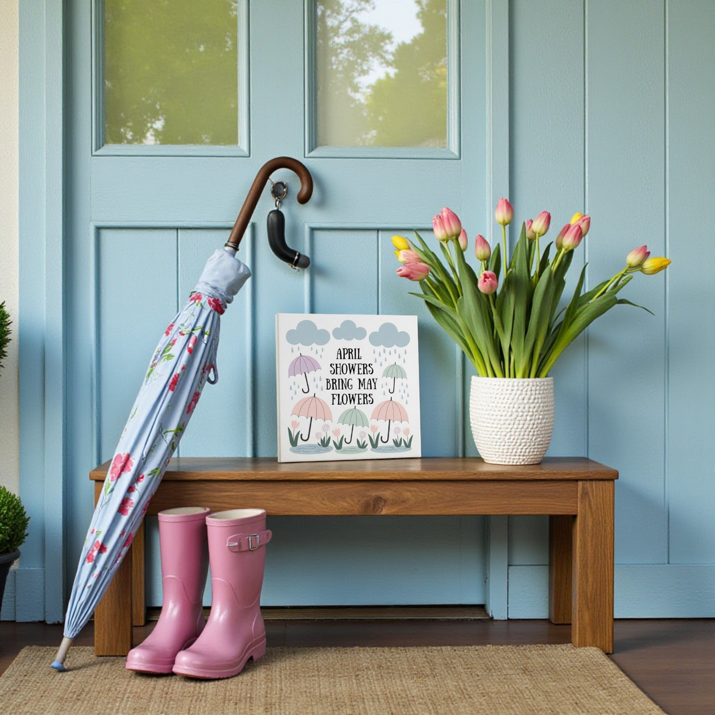 The April Showers Bring May Flowers Canvas Art by Mirabilia Boutique features a wooden bench with pink tulips, rain boots, and a floral umbrella, set against a blue door—perfect spring wall art or nursery decor.