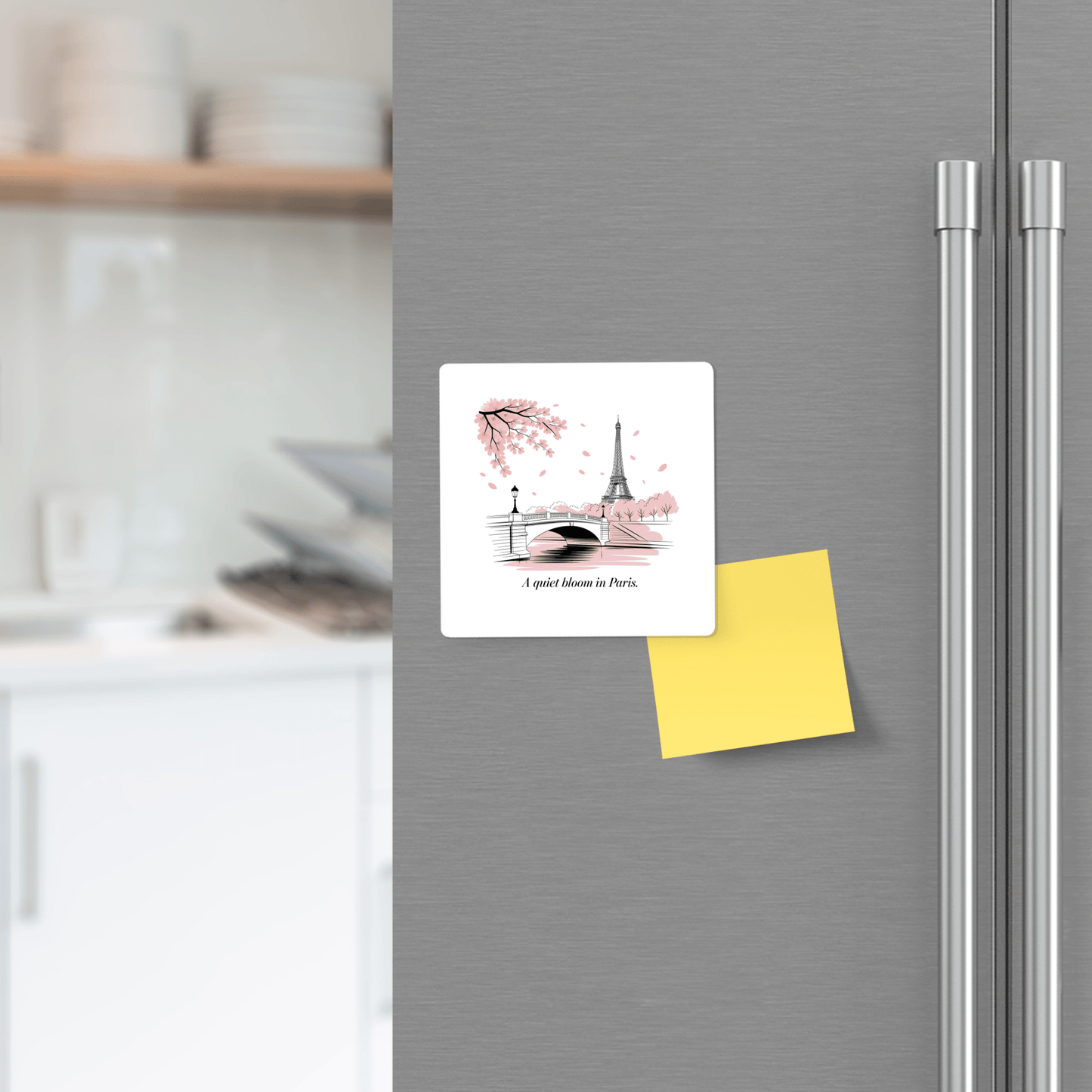 A refrigerator door features Mirabilia Boutique’s “A Quiet Bloom in Paris – Cherry Blossom Magnet” and a blank yellow sticky note, with blurred kitchen shelves and dishes in the background.