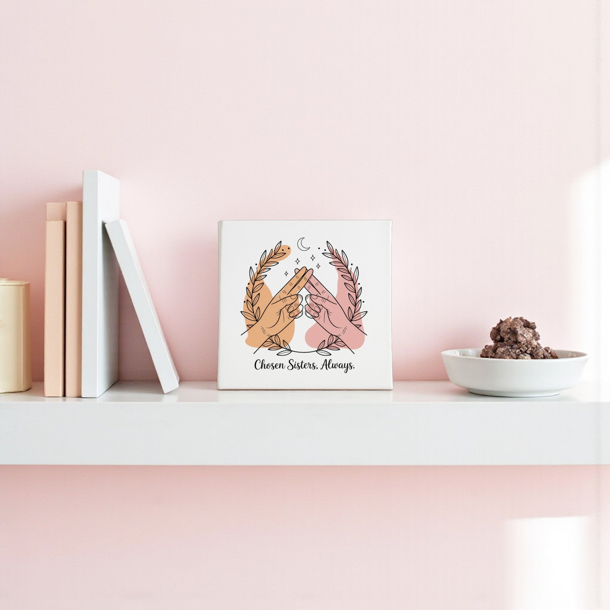 A white shelf on a pink wall holds books, cookies, and the Mirabilia Boutique Chosen Sisters Canvas, featuring two hands high-fiving beneath laurel branches and stars. The canvas reads, "Chosen Sisters. Always."—a perfect friendship gift.