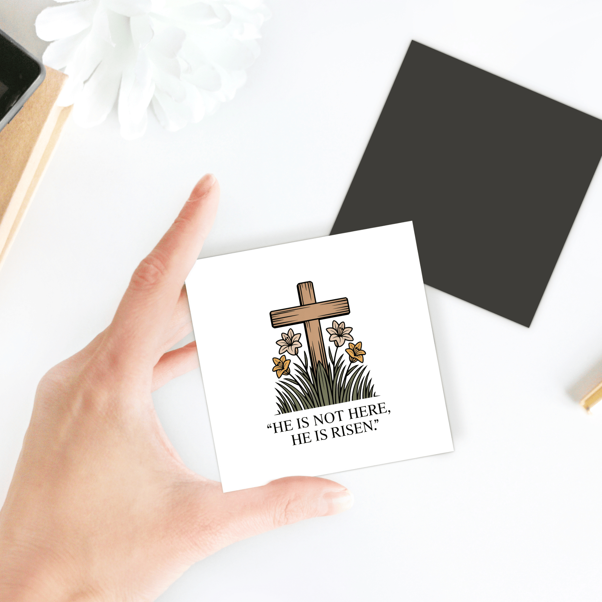 A hand holds the "He Is Risen Easter Magnet – Cross & Lily Scripture Art" by Mirabilia Boutique, featuring a wooden cross, lilies, and the words "HE IS NOT HERE, HE IS RISEN." A black scripture magnet and white flower are in the background.