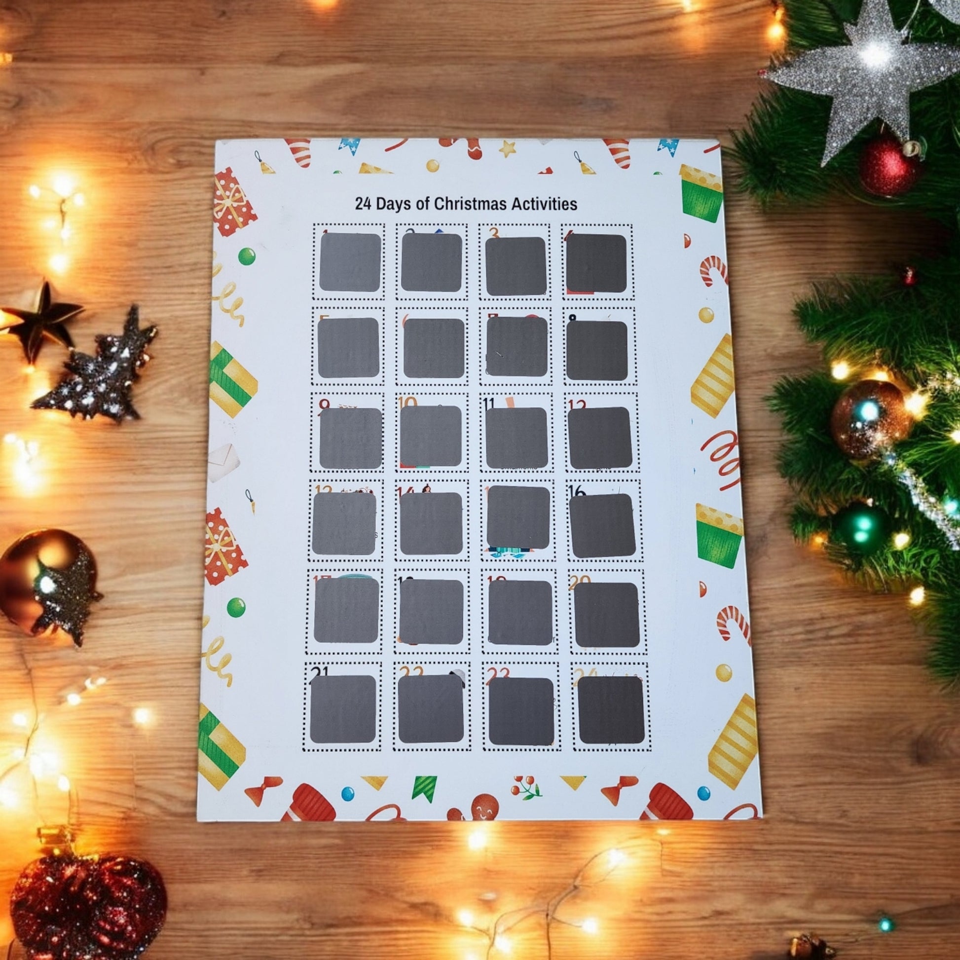 A 24 Days of Christmas Activities Scratch Off Calendar by Mirabilia Boutique is displayed on a wooden table, nestled among festive decorations like star ornaments, baubles, and a pine branch. The calendar features numbered squares framed with holiday-themed illustrations, adding a playful touch to your holiday décor.