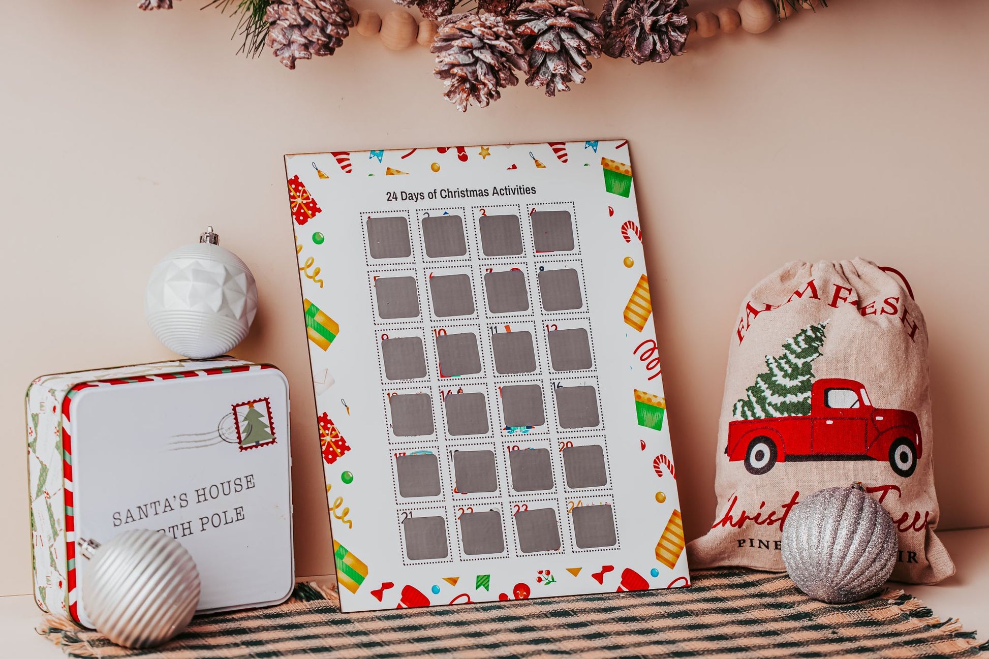 A festive scene features the Mirabilia Boutique Scratch-Off Christmas Activity Calendar—Holiday Countdown for Kids with 25 family activities, a "Santa's House North Pole" tin, red truck sack, ornaments, and pinecones on a checkered cloth.