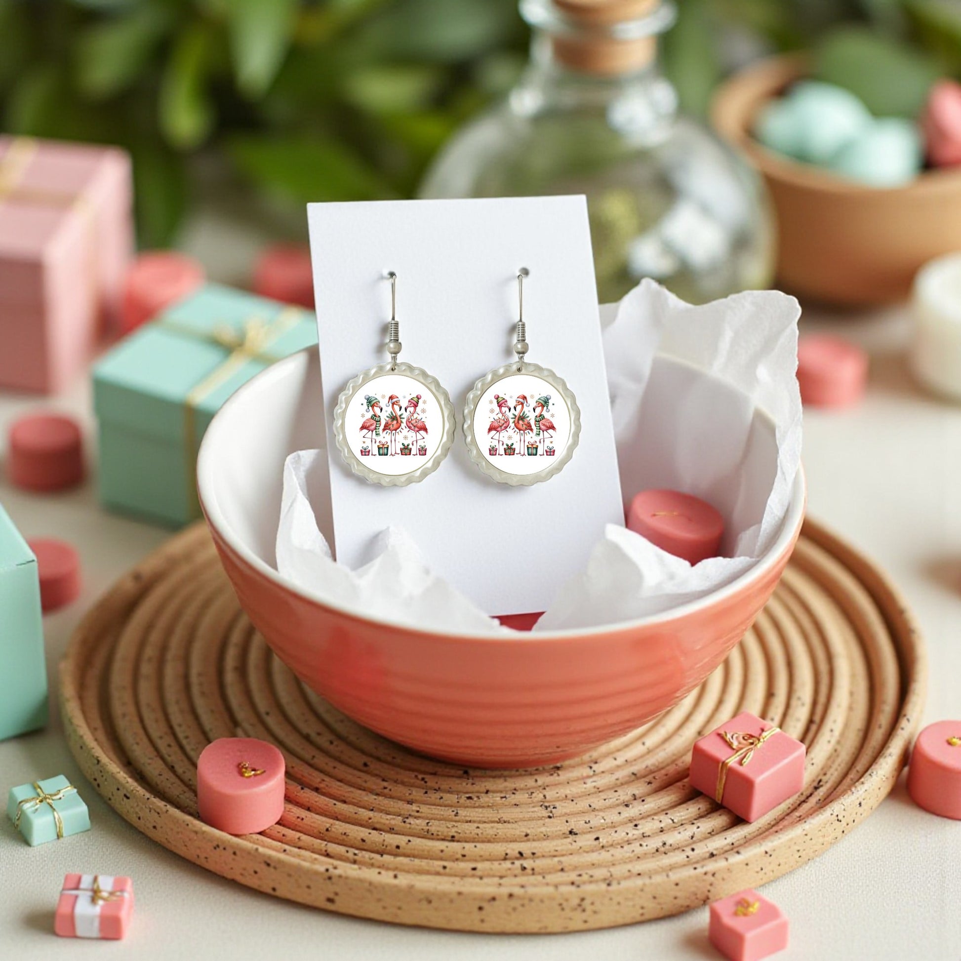 The Christmas Flamingo Earrings by Mirabilia Boutique, vibrant and hypoallergenic, are displayed on a card in a pale pink bowl with tissue and surrounded by festive gift boxes—perfect for gifting tropical holiday jewelry.