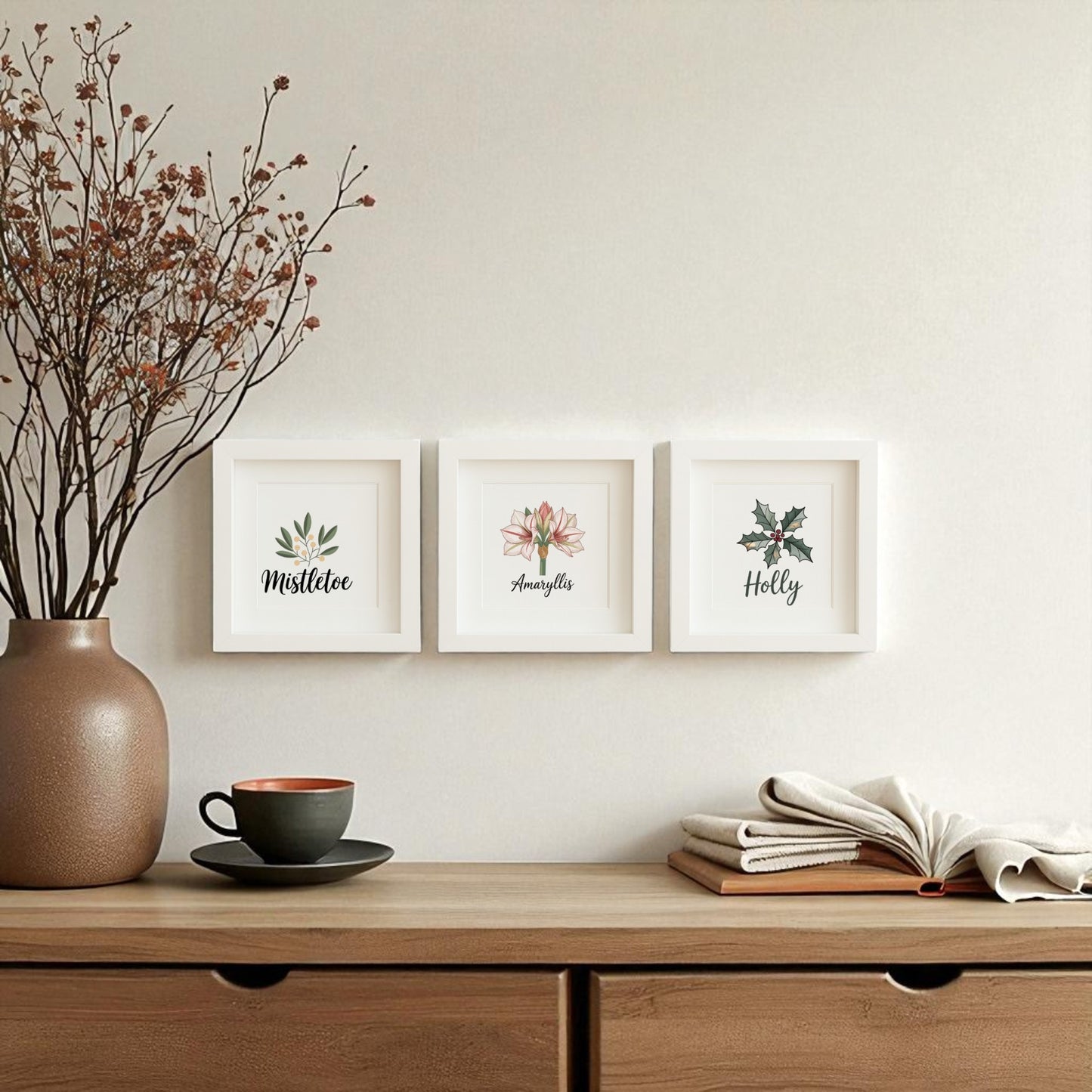 A wooden sideboard holds a brown vase with dried branches, a cup and saucer, and open books. Above, Mirabilia Boutique's "Mistletoe Amaryllis Holly Prints | Holiday Shadowbox Trio" decorates the cream wall.