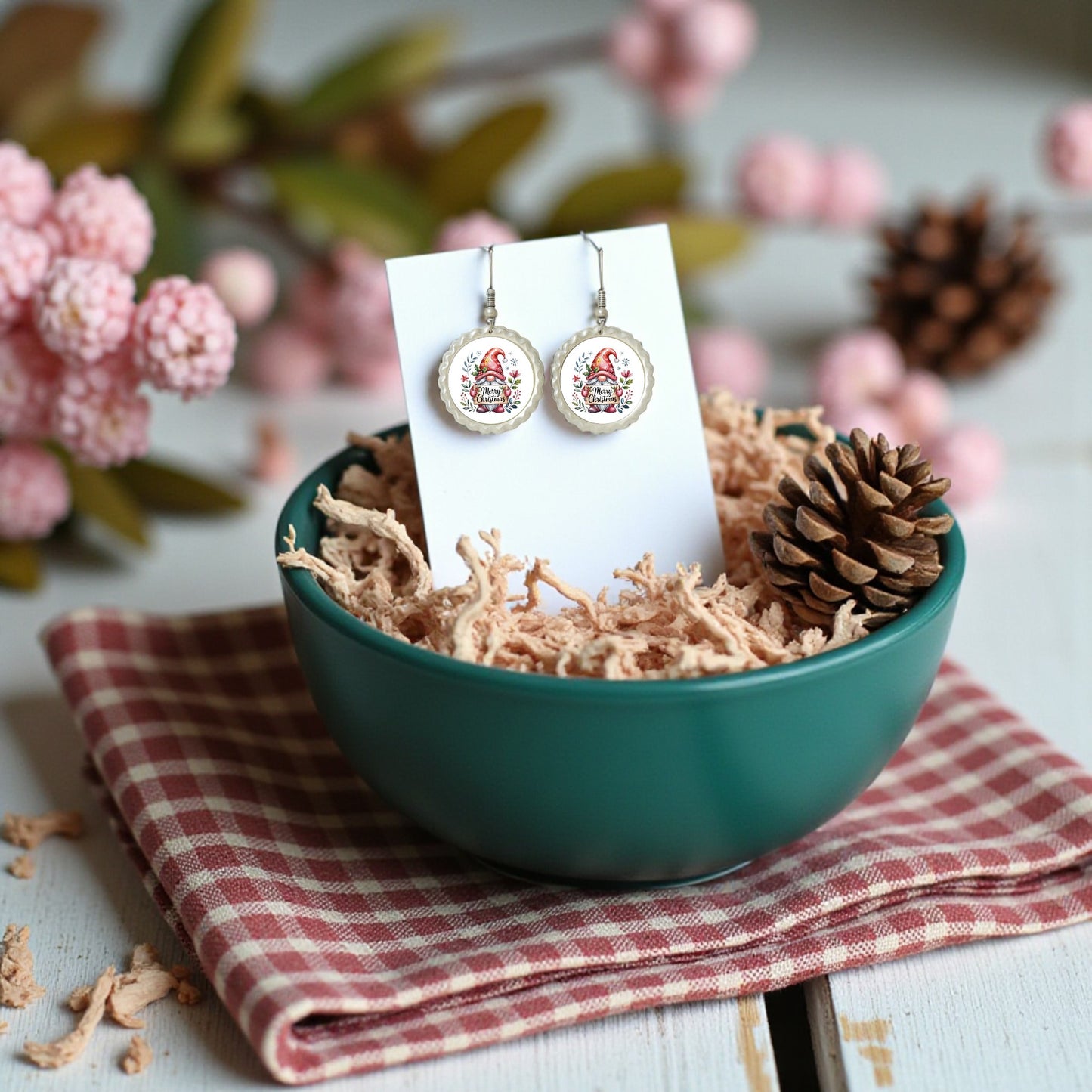 Mirabilia Boutique's Merry Christmas Gnome Earrings—festive holiday jewelry—are displayed on a card in a teal bowl with straw, beside a pinecone and red checked napkin, with pink flowers and pinecones in the background.
