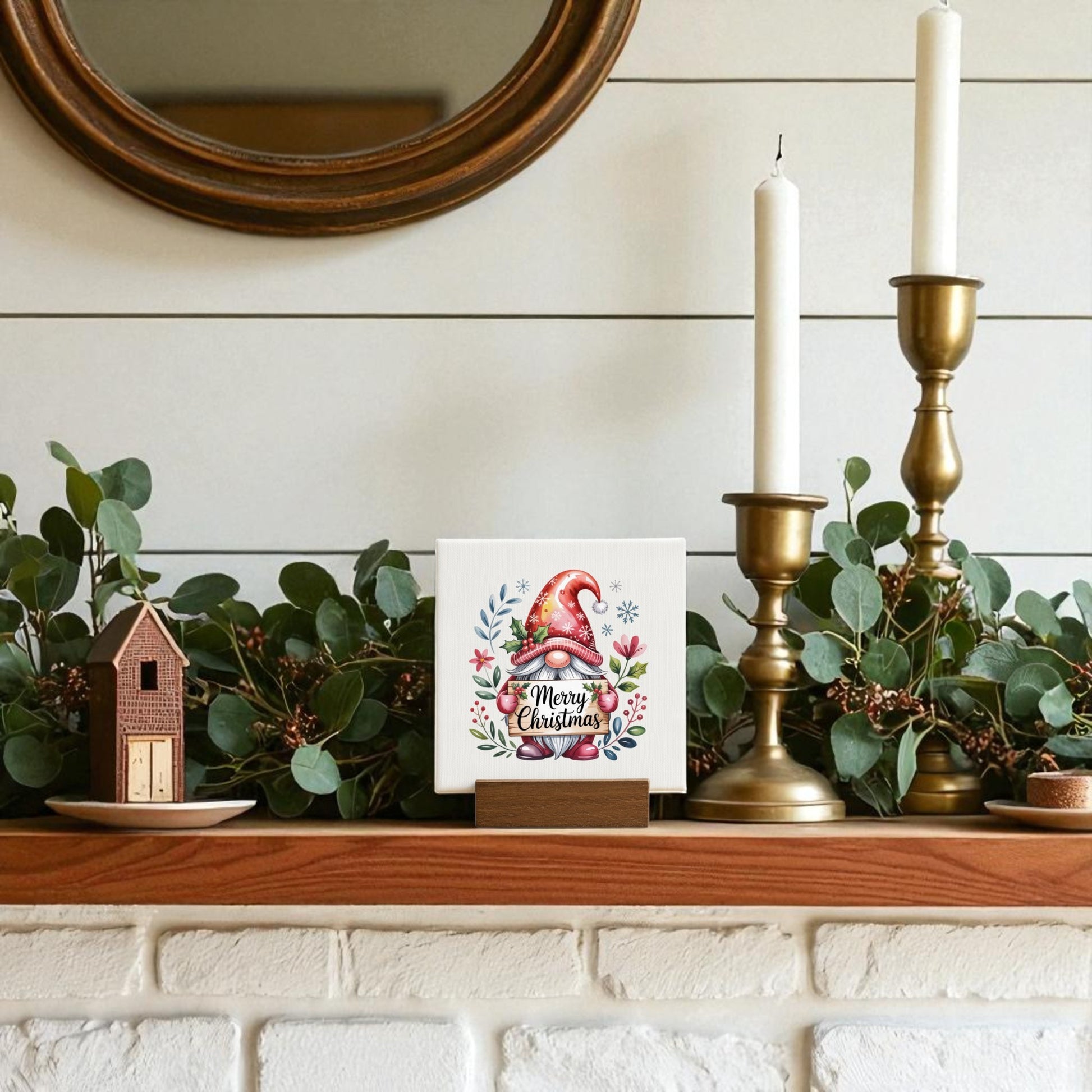 A white shiplap mantel is adorned with greenery, brass candlesticks, a small house ornament, and the Mirabilia Boutique Christmas Gnome Mini Sign (4x4 Canvas)—a festive touch beneath a round mirror for holiday home decor.