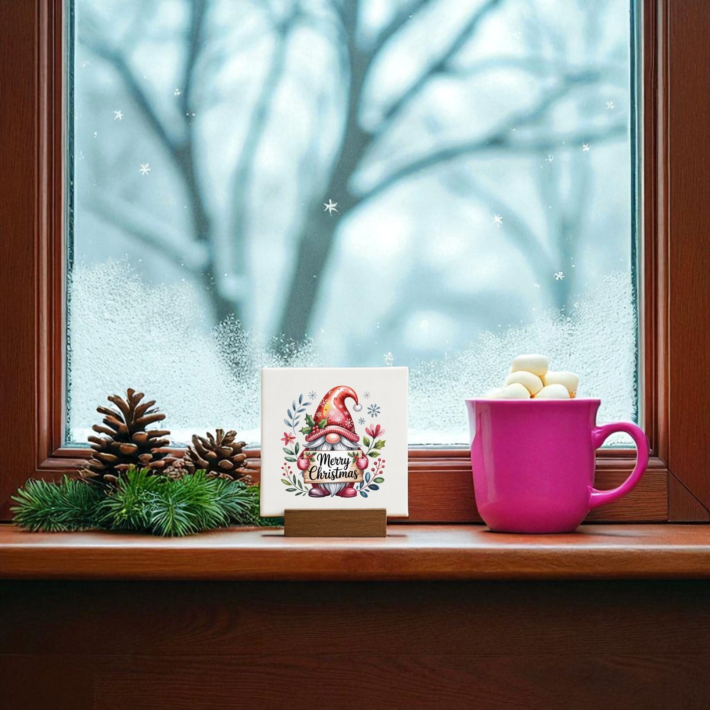A pink mug with marshmallows, pinecones, greenery, and a “Christmas Gnome Mini Sign” 4x4 canvas decor by Mirabilia Boutique sit on a wooden windowsill. Snowy trees are visible outside through the frosted window.