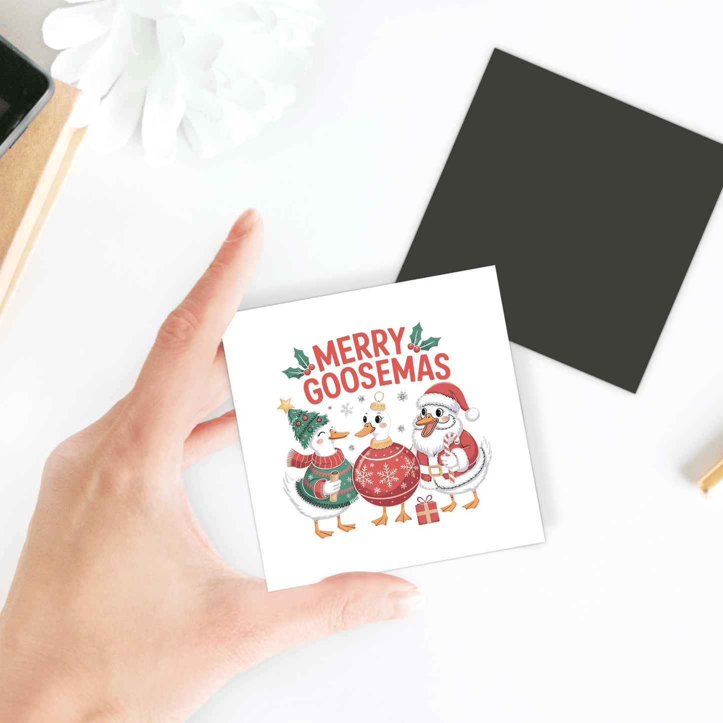 A hand holds the "Merry Goosemas Magnet" by Mirabilia Boutique featuring three festive geese in winter hats and scarves under the words "Merry Goosemas"—a funny Christmas goose gift or quirky holiday fridge decor. A black square and white flower are nearby.