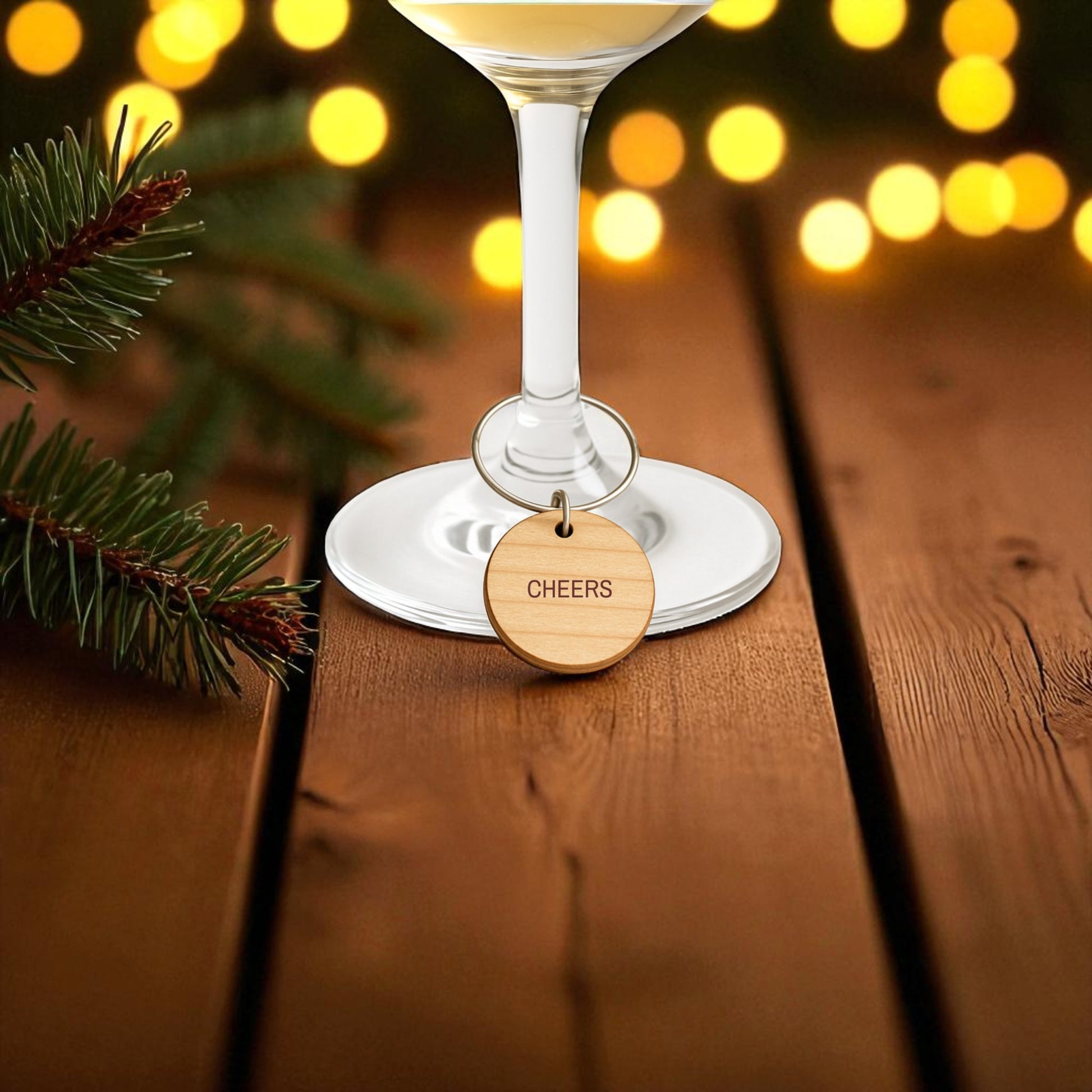 A wine glass with a wooden "CHEERS" charm from the Mirabilia Boutique Christmas Wine Charms | Wood Holiday Glass Markers Set sits on a wooden table with pine branches and warm holiday lights softly glowing in the background.