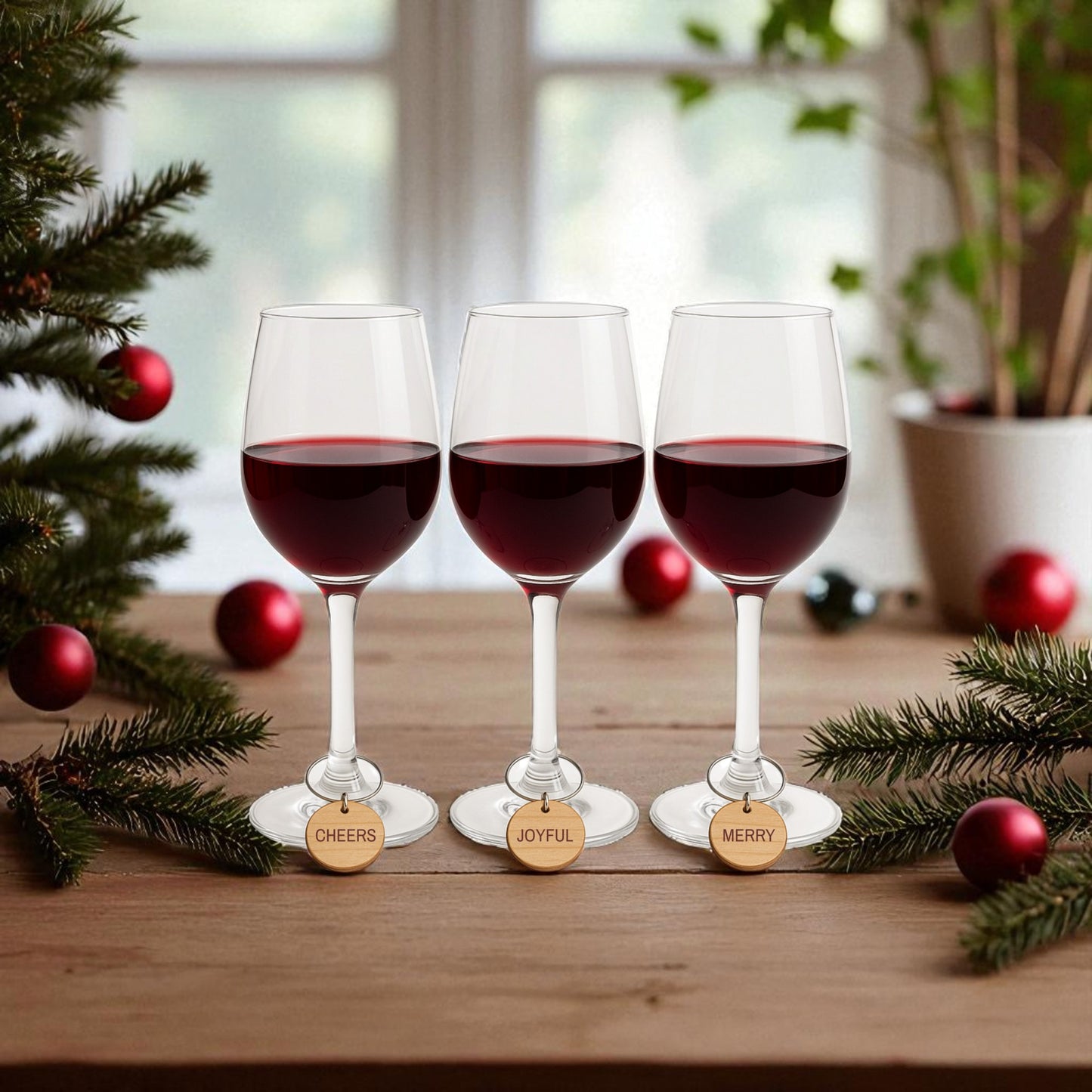 Three red wine glasses on a festive table display Mirabilia Boutique’s Christmas Wine Charms—wooden holiday glass markers reading "CHEERS," "JOYFUL," and "MERRY"—adding charm to your holiday entertaining. A window is visible in the background.