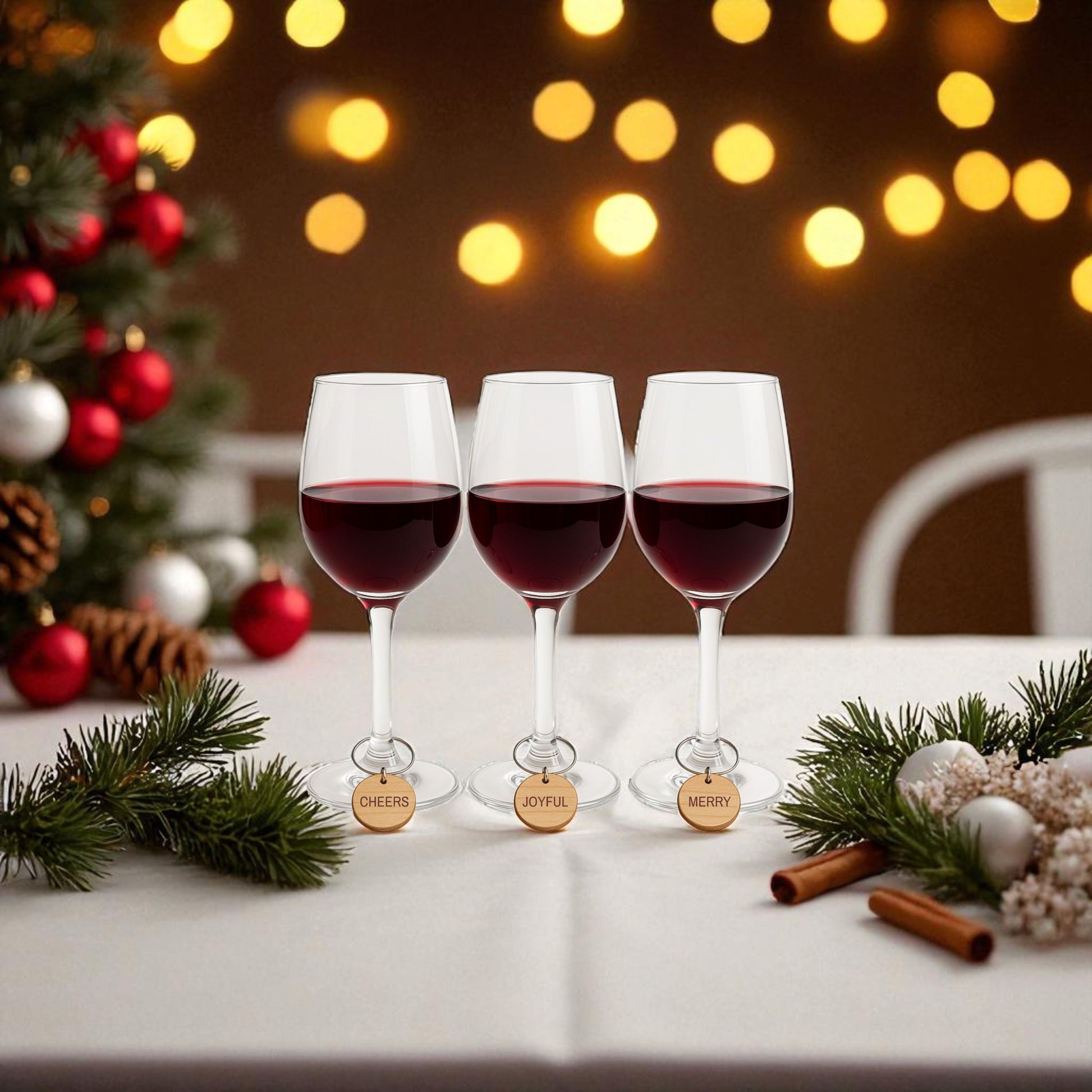 Three glasses with Mirabilia Boutique's Christmas Wine Charms | Wood Holiday Glass Markers Set sit on a festive table with pine and ornaments, blurred holiday lights, and a glowing Christmas tree in the background.