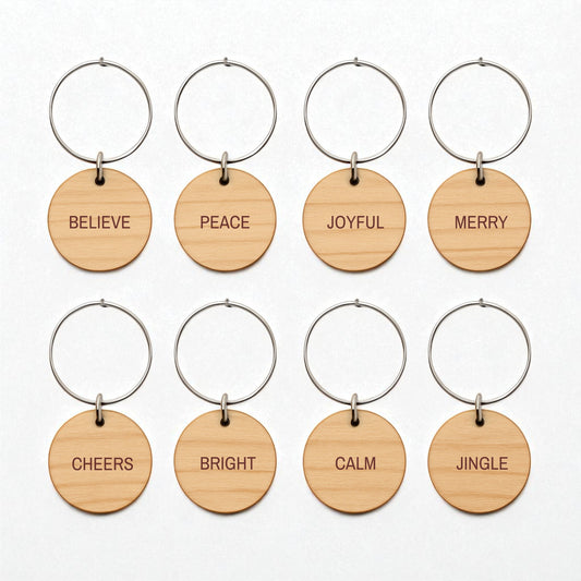 Mirabilia Boutique’s Christmas Wine Charms | Wood Holiday Glass Markers Set includes eight round wooden keychains with metal rings, each engraved with a festive word—perfect for marking glasses at your holiday gatherings.