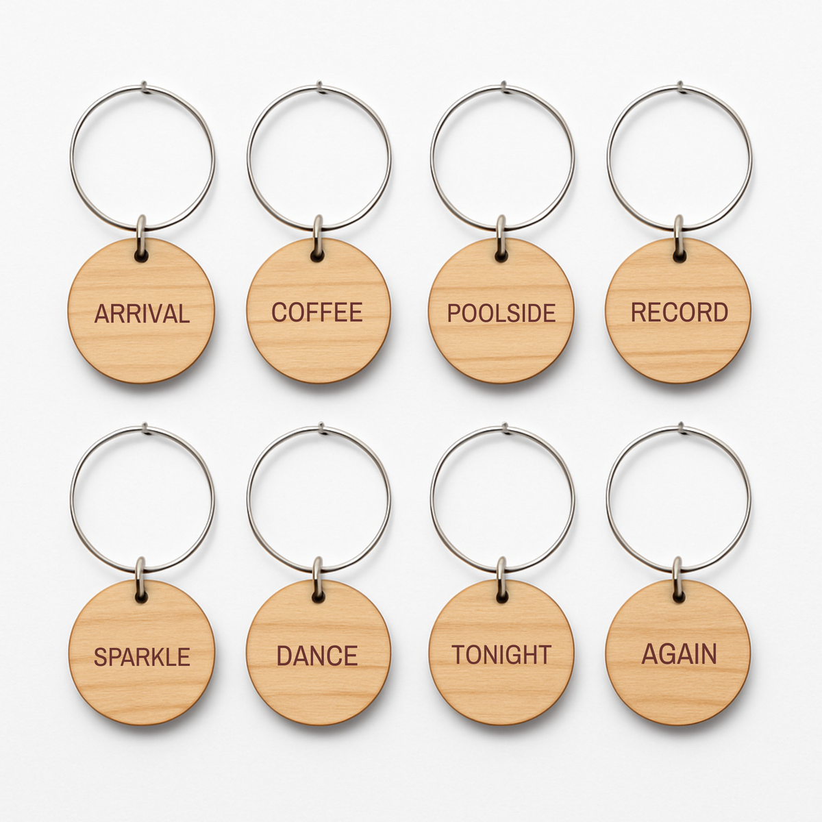Eight circular wooden Chrome & Starlight Wood Wine Charms on silver metal rings, each engraved with a unique word: “Arrival,” “Coffee,” “Poolside,” “Record,” “Sparkle,” “Dance,” “Tonight,” and “Again.”.