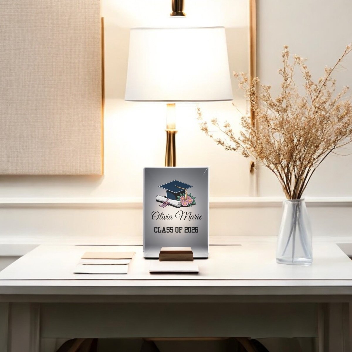 A Mirabilia Boutique Floral Personalized Graduation Night Light—featuring dried flowers, a custom “Olivia Marie, Class of 2026” plaque with cap and diploma—sits in a softly lit, neutral room: the perfect graduation gift.