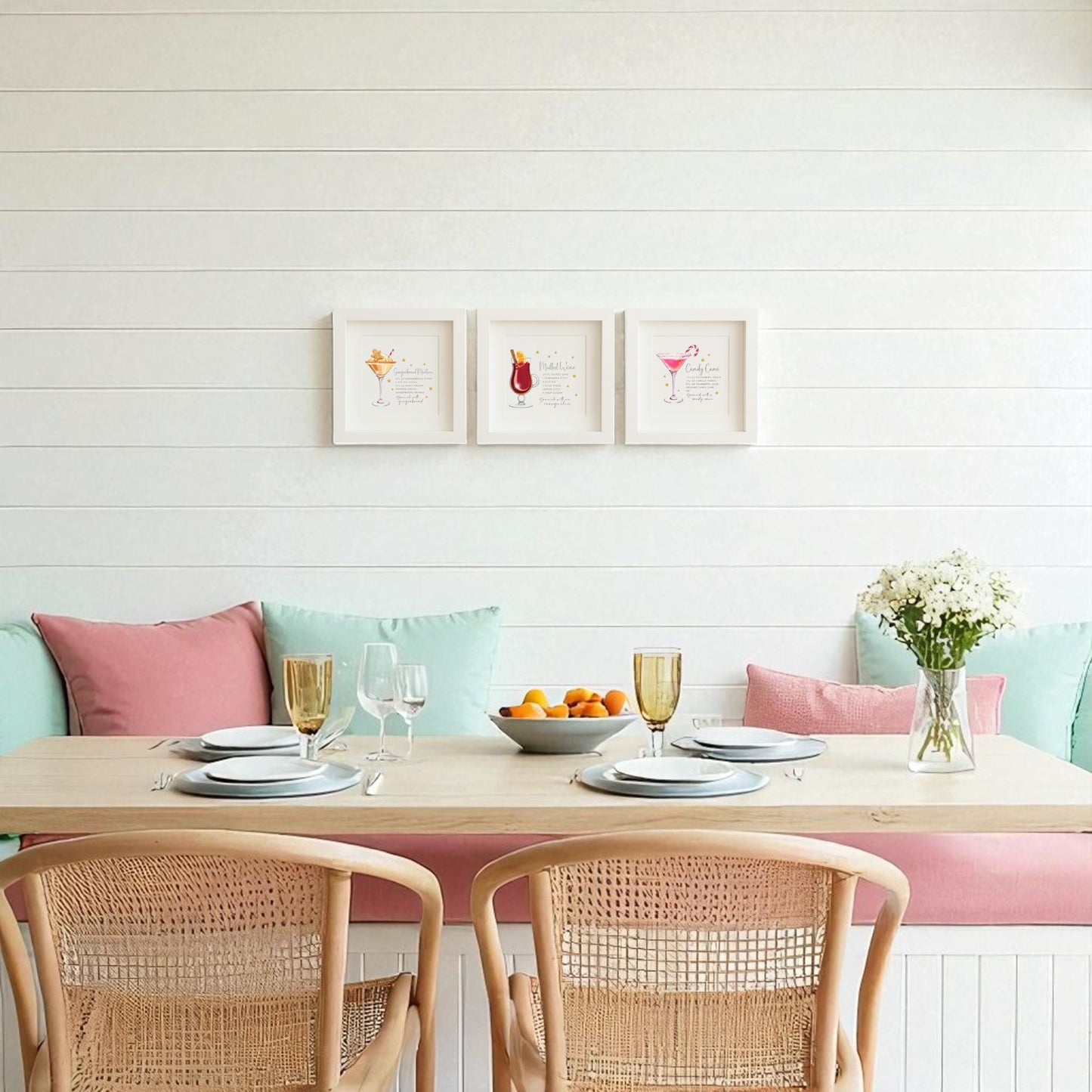 A bright dining area with a wooden table for four, pastel cushions, rattan chairs, a vase of white flowers, and the Mirabilia Boutique Classic Christmas Cocktails 5x5 Shadowbox Trio holiday bar art on the white shiplap wall.