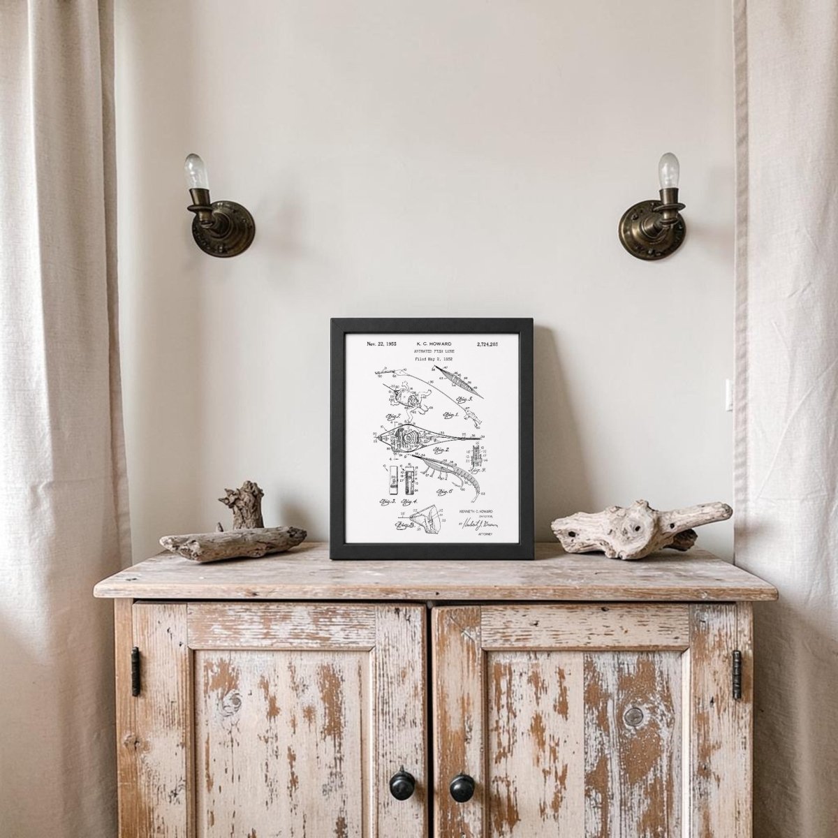 A rustic wooden cabinet with a distressed finish stands against a beige wall. On top sits the Mirabilia Boutique Animated Fish Lure Patent Art Print, accompanied by two pieces of driftwood and exposed-bulb sconces above.