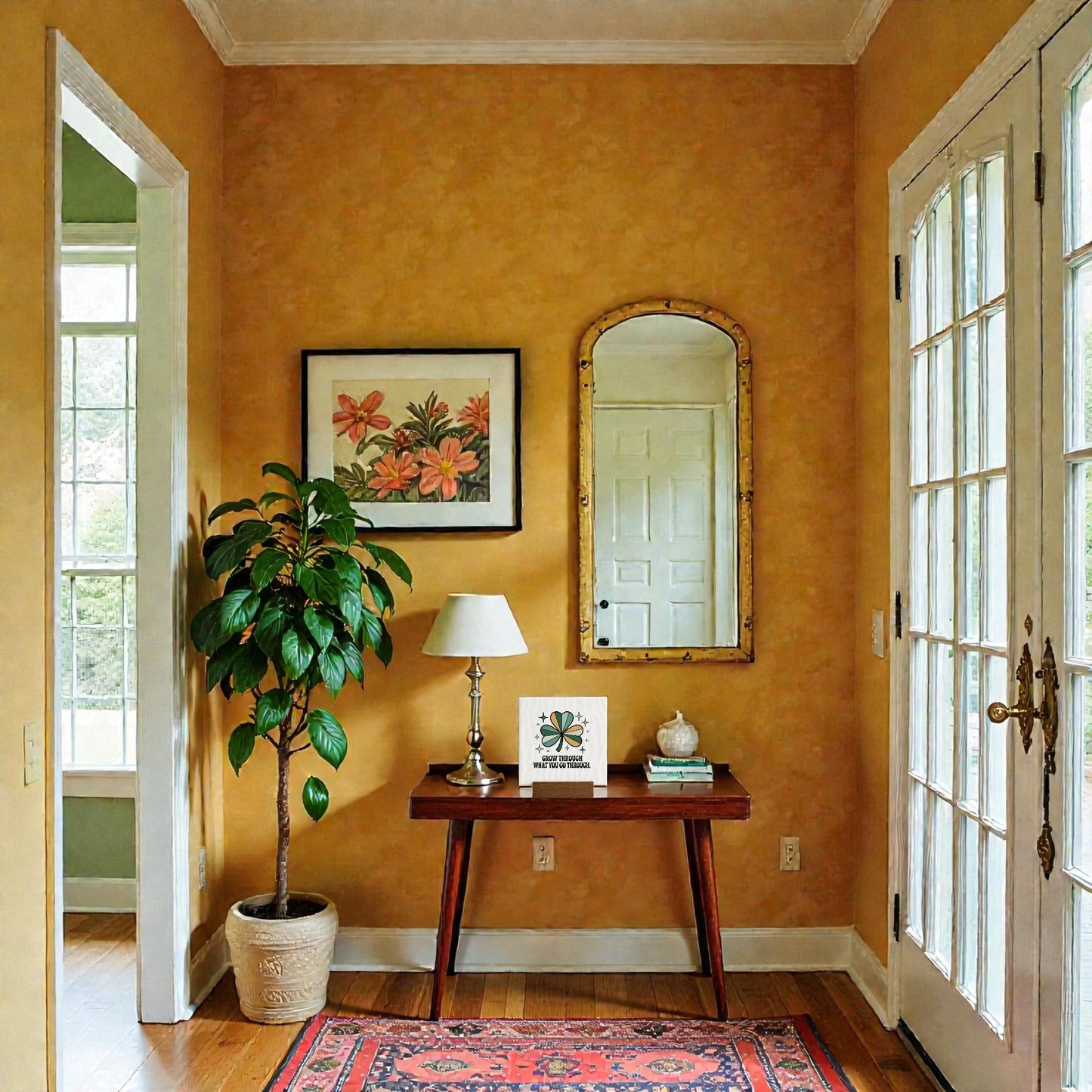 A cozy entryway features mustard yellow walls, a framed flower print, gold-framed mirror, four-leaf clover decor, and a wooden table with a lamp, books, Mirabilia Boutique’s "Grow Through What You Go Through" mini sign, and a small dish. Sunlight streams in.