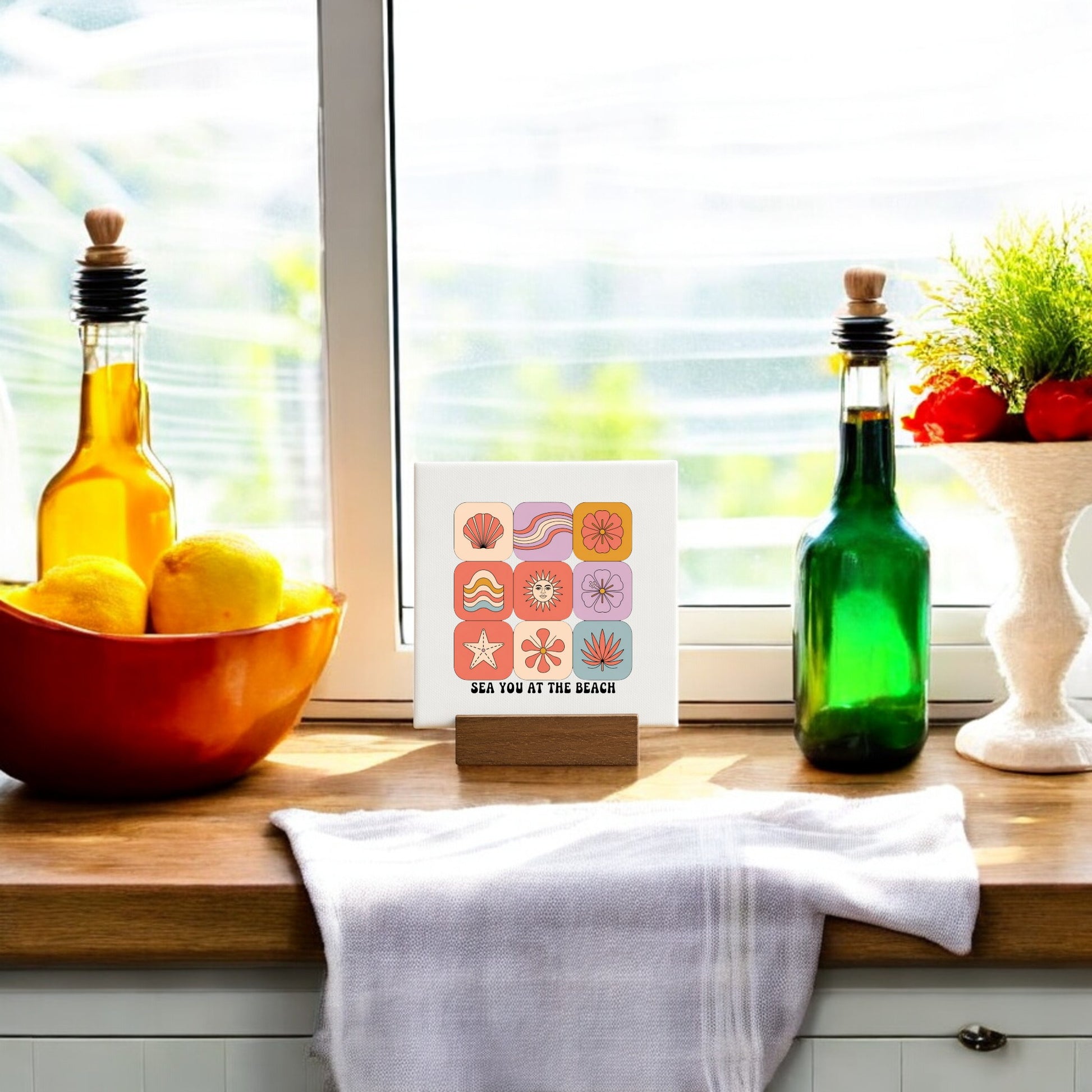 A kitchen windowsill with oil and vinegar bottles, lemons, a white towel, and the Mirabilia Boutique "Sea You at the Beach Mini Canvas – Coastal Summer Vibes”—a charming touch of coastal decor.