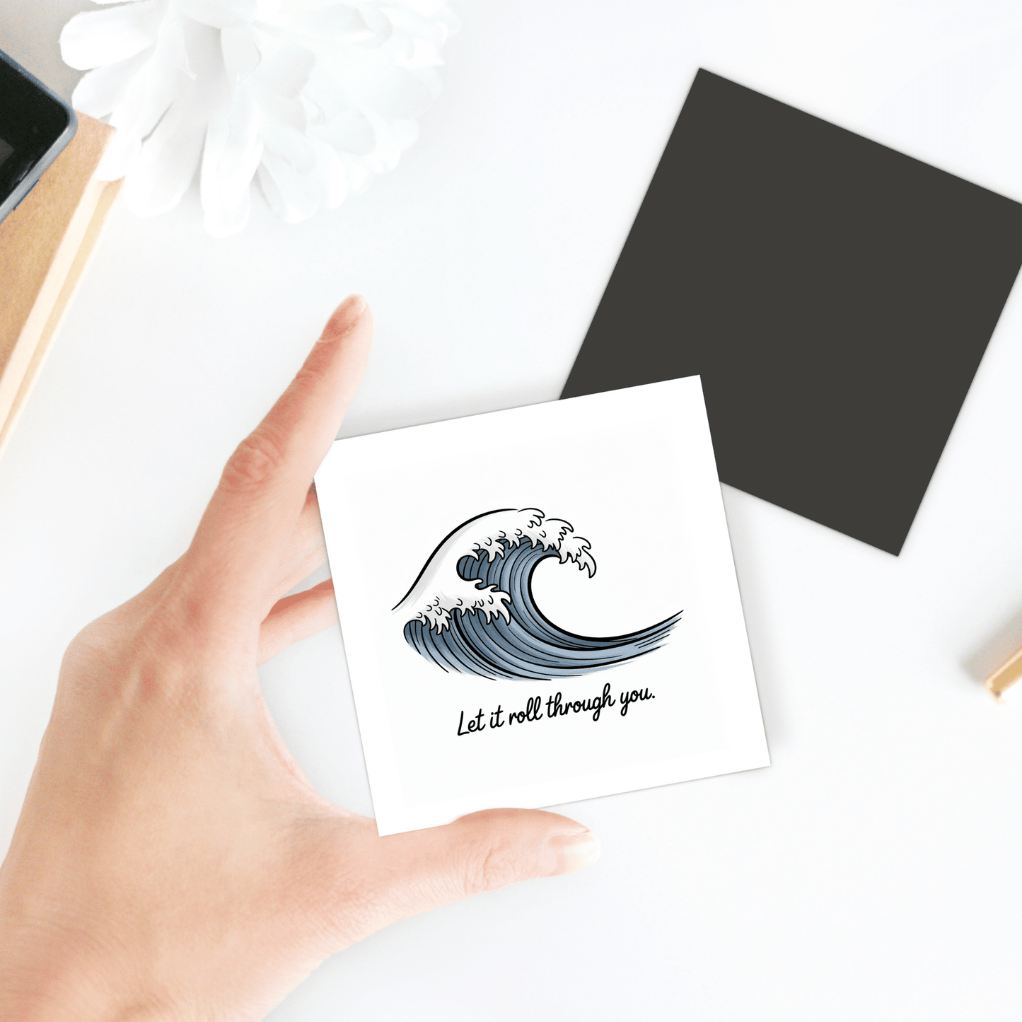 A hand holds the Let It Roll Through You Magnet – Ocean-Inspired Calm by Mirabilia Boutique, featuring a blue wave and calming message. In the background, a black square and white flower sit on a white surface.
