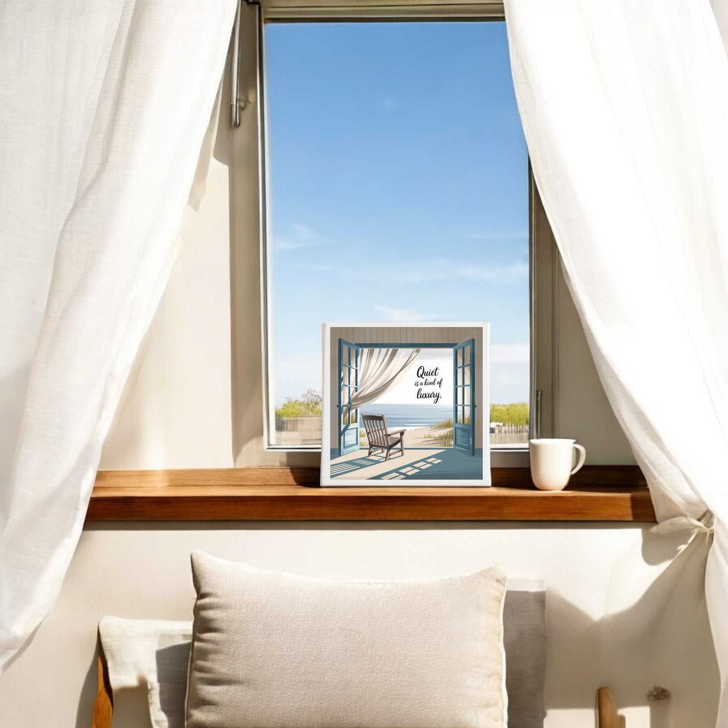 A cozy window seat with white cushions and a mug sits beneath an open window with sheer curtains. On the windowsill is Mirabilia Boutique's Quiet Is a Kind of Luxury Canvas, adding peaceful summer wall art and quiet luxury decor to the scene. Blue sky is visible outside.