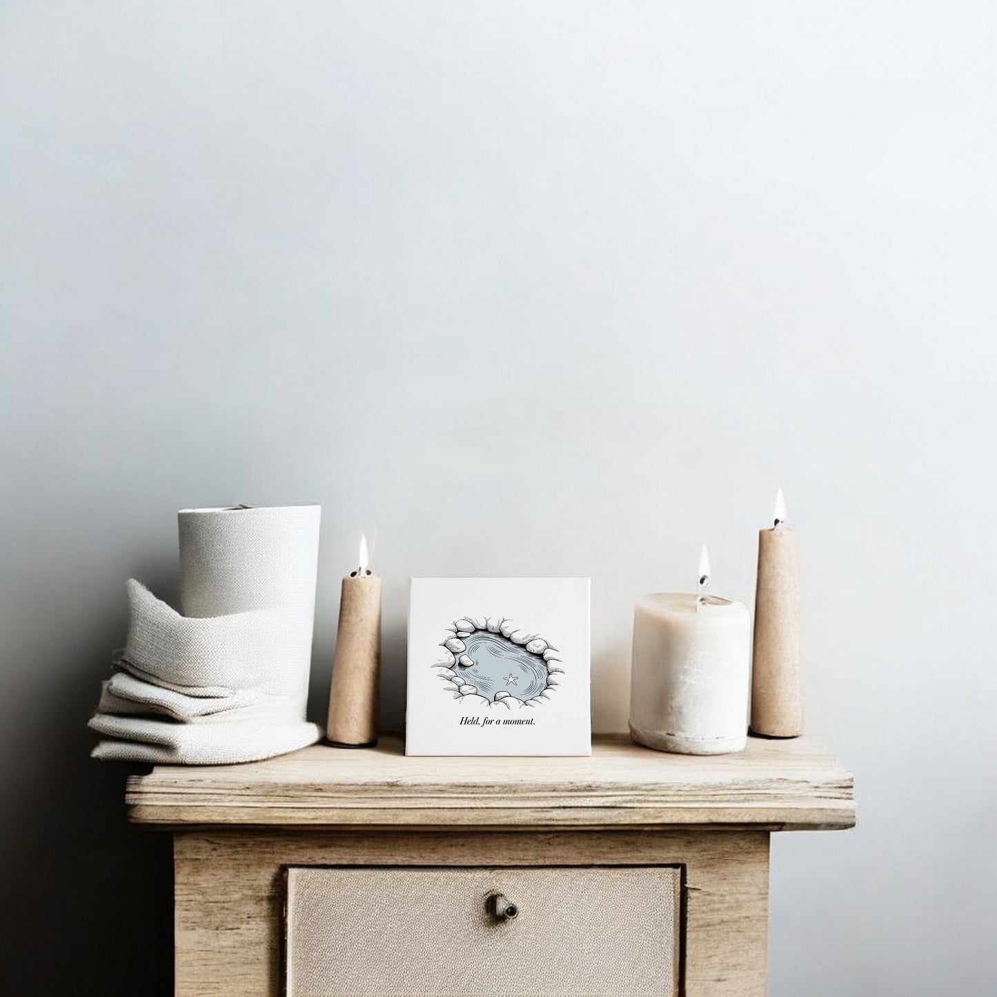 A small wooden table holds folded towels, three lit candles, and a card beside Mirabilia Boutique's "Held, For a Moment" 8x8 Coastal Canvas Wall Art on a plain light gray wall.