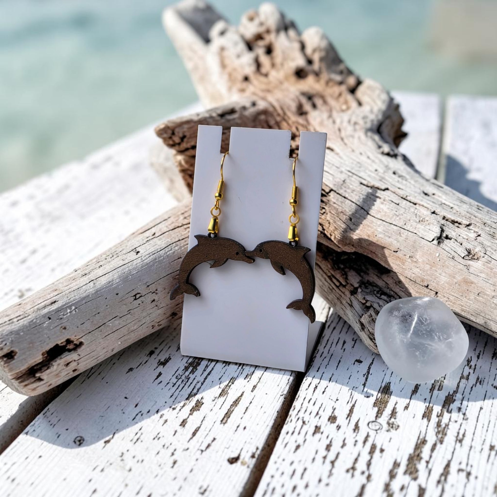 Mirabilia Boutique's Dolphin Earrings, lasercut from walnut wood, hang from gold hooks on a white card displayed with driftwood and glass pebbles on weathered wood, evoking ocean beauty against a blurred beach backdrop.